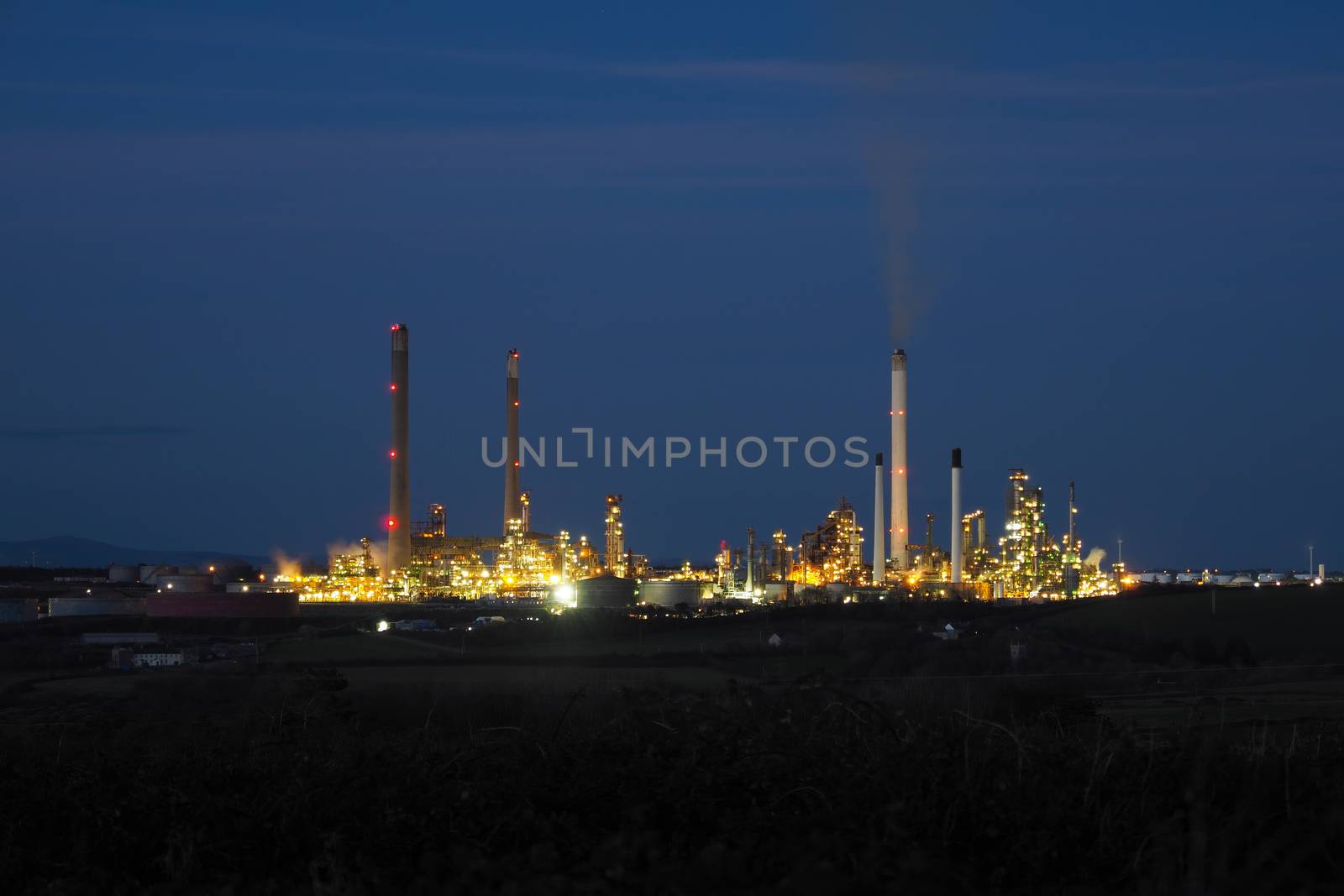Pembroke oil refinery lit up at night with smoke rising from a chimney stack, Rhoscrowther on the Pembrokeshire coast, near Milford Haven, Wales, UK