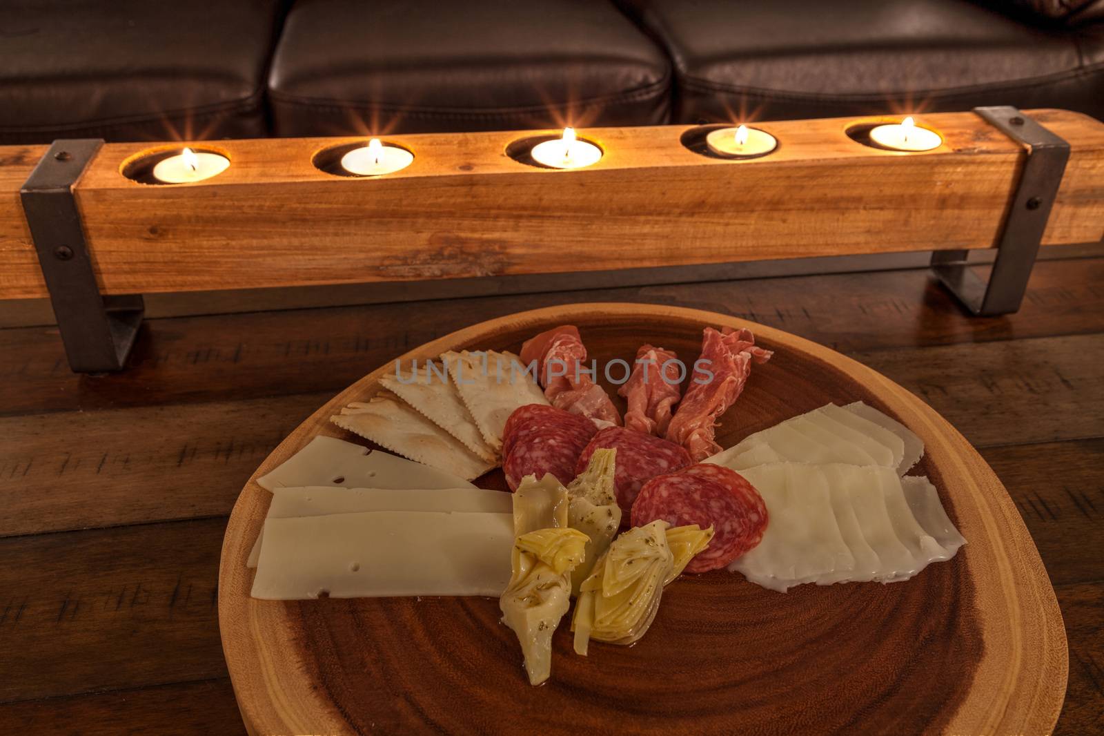 Charcuterie board on rustic wood with candles behind a spread of prosciutto, mozzarella cheese, Genoa salami, Fontina cheese and artisanal crackers.