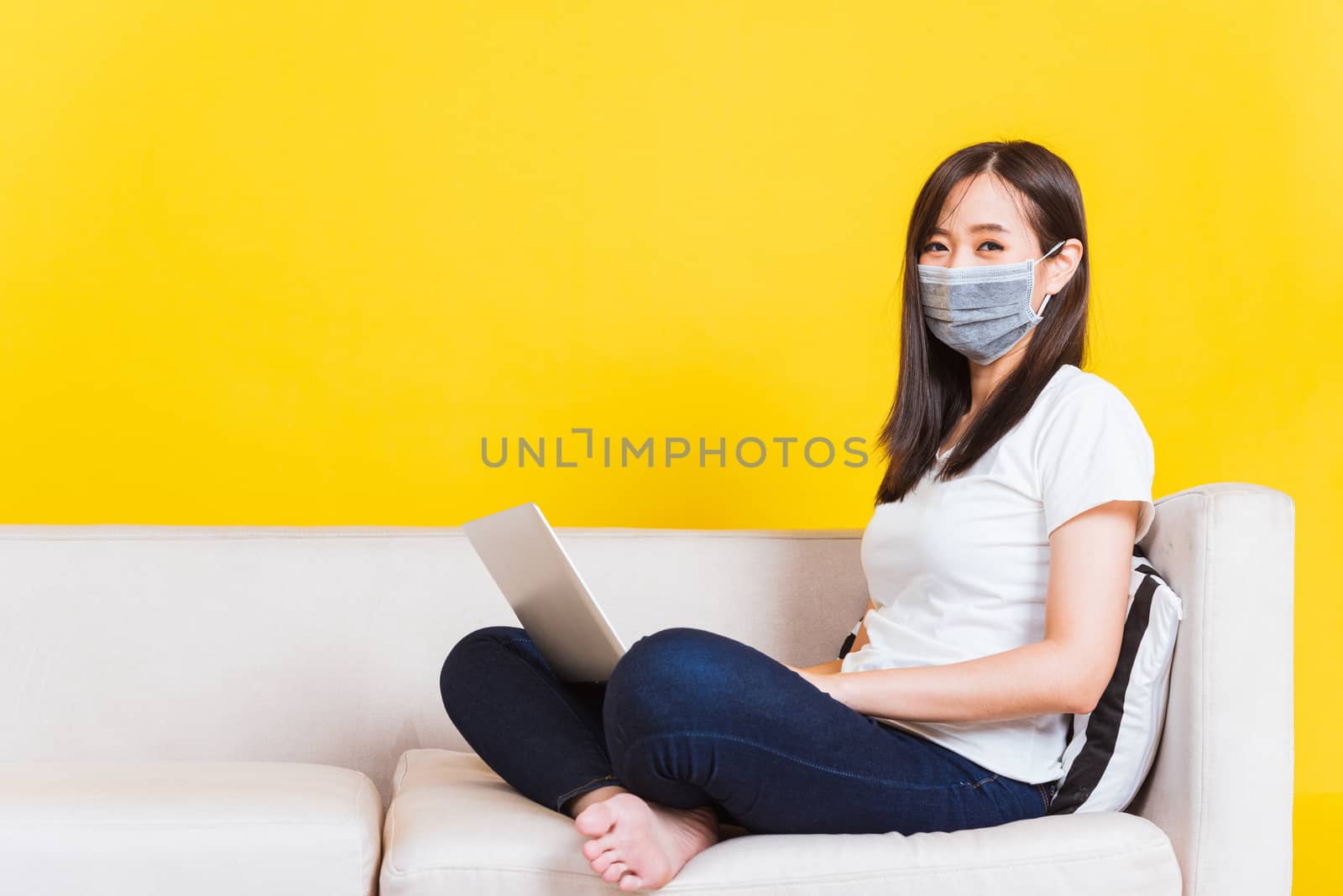 Portrait Asian of beautiful young woman sitting on sofa wearing medical face mask protective she work from home with laptop computer during Coronavirus studio shot isolated on yellow background