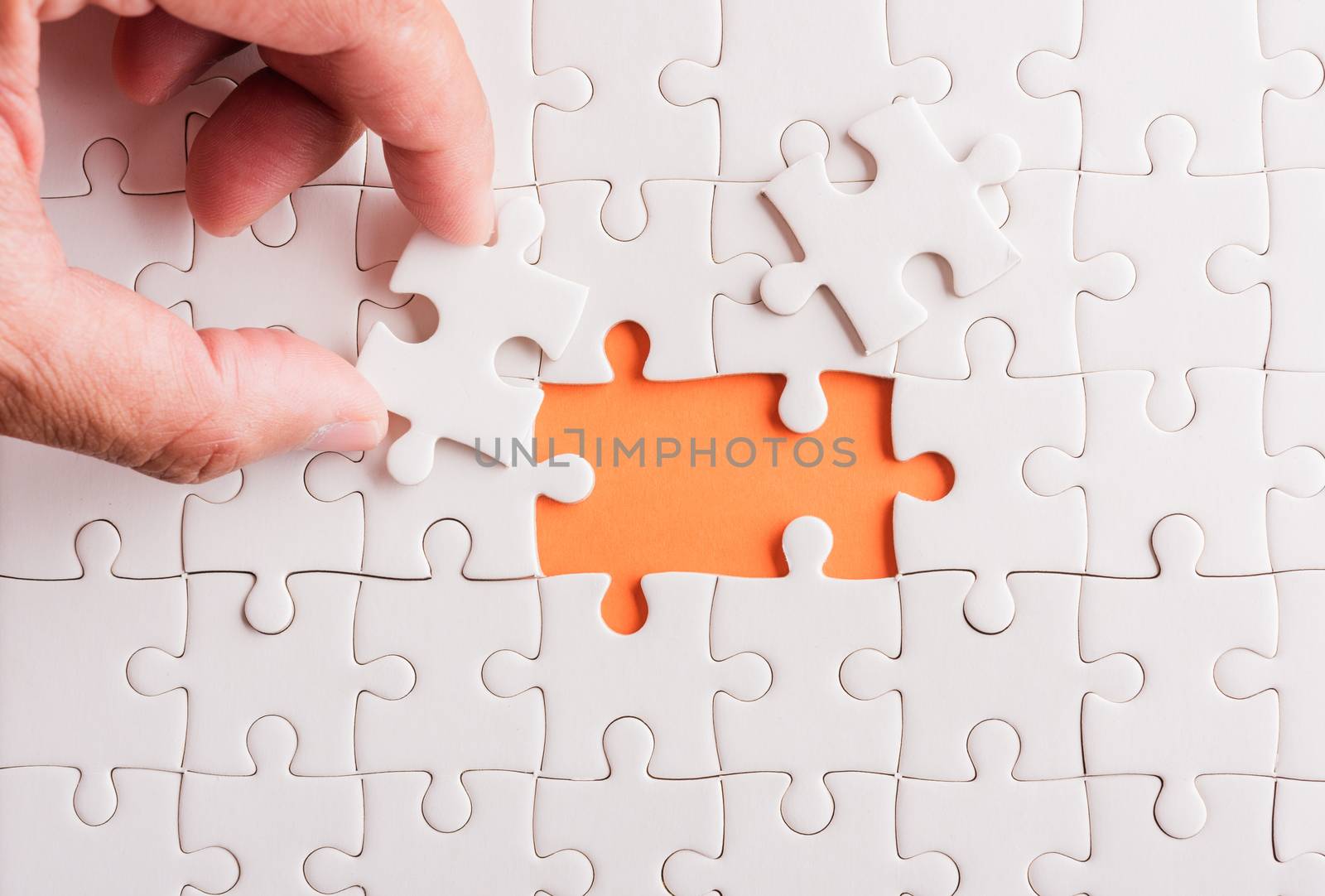 Top view flat lay of hand-holding last piece white paper jigsaw puzzle game last pieces put to place for solve problem complete mission, studio shot on an orange background, quiz calculation concept