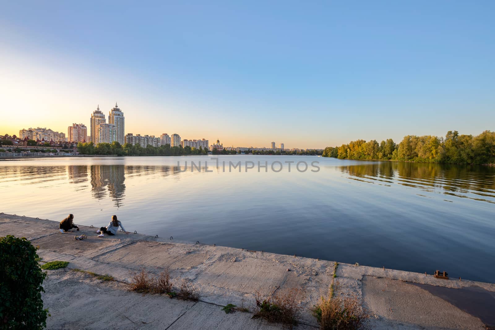 Kiev / Ukraine - September 22, 2020 - Two girls on the embankment close to the high Obolon buildings near the Dnieper river in Kiev, Ukraine. Blue clear sky and reflection in the water.