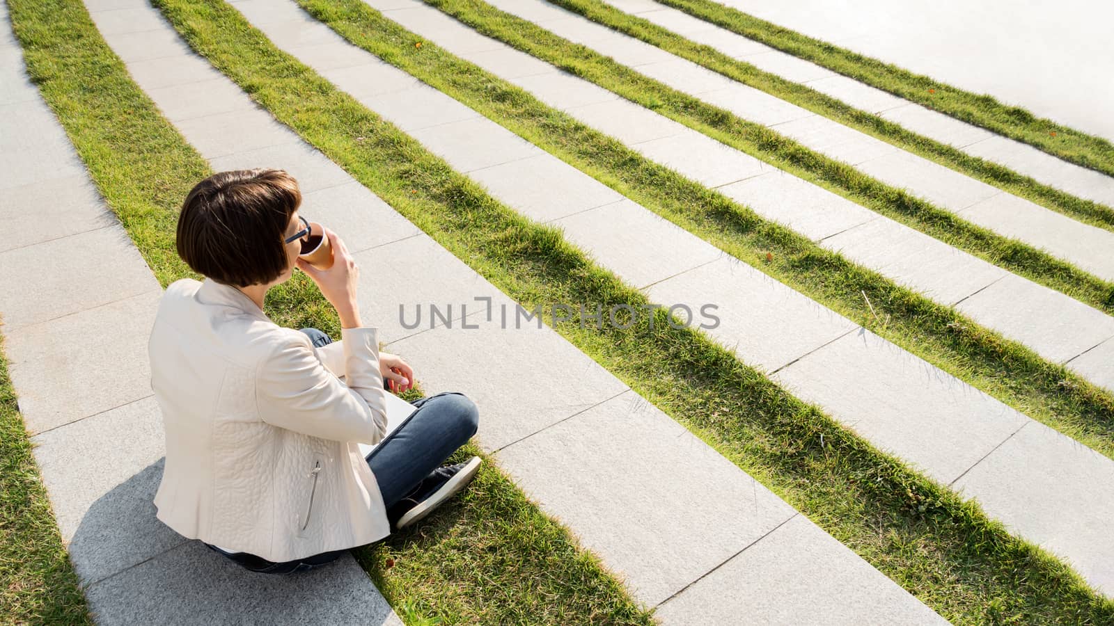 Woman with short haircut and eyeglasses sits in park with take a by aksenovko