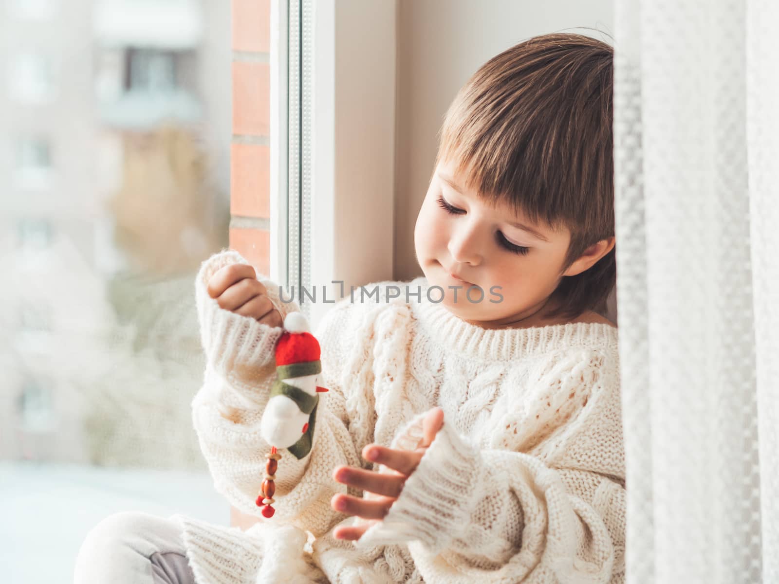 Kid with felt decorative snowman for Christmas tree. Boy in cable-knit oversized sweater. Cozy outfit for snuggle weather. Funny character with red Santa Claus hat. Winter holiday spirit. New year.