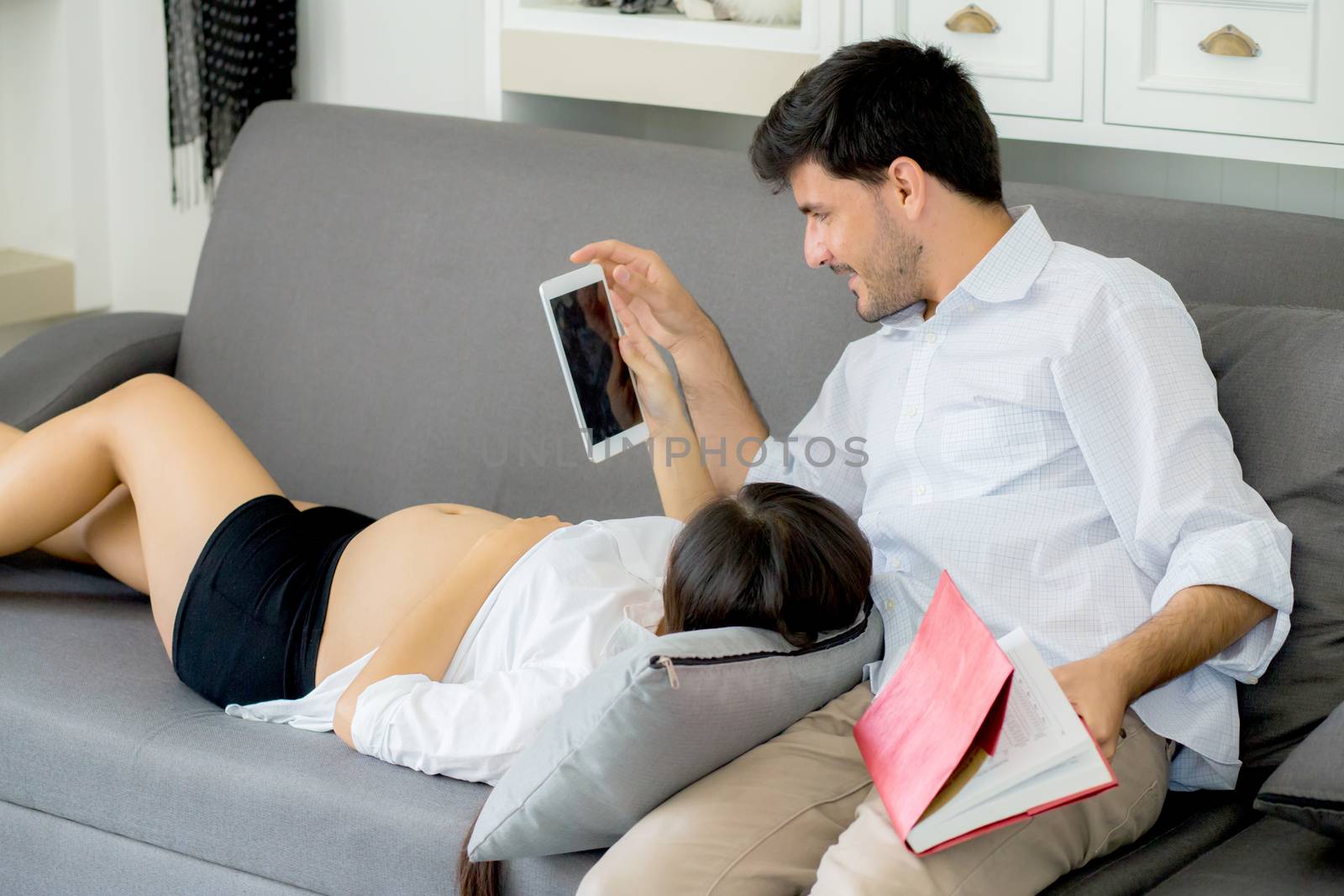 Asian couple family young mother and husband using a tablet at home, woman and man is pregnant lying on sofa looking tablet read book.