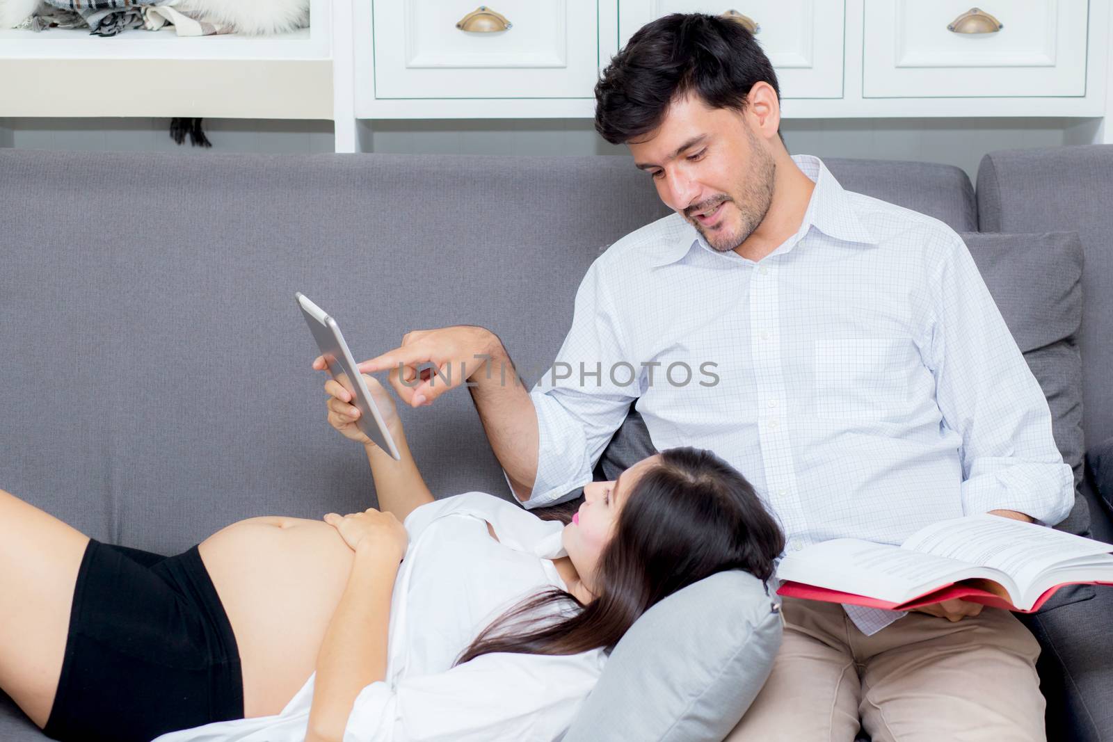 Asian couple family young mother and husband using a tablet at home, woman and man is pregnant lying on sofa looking tablet read book.