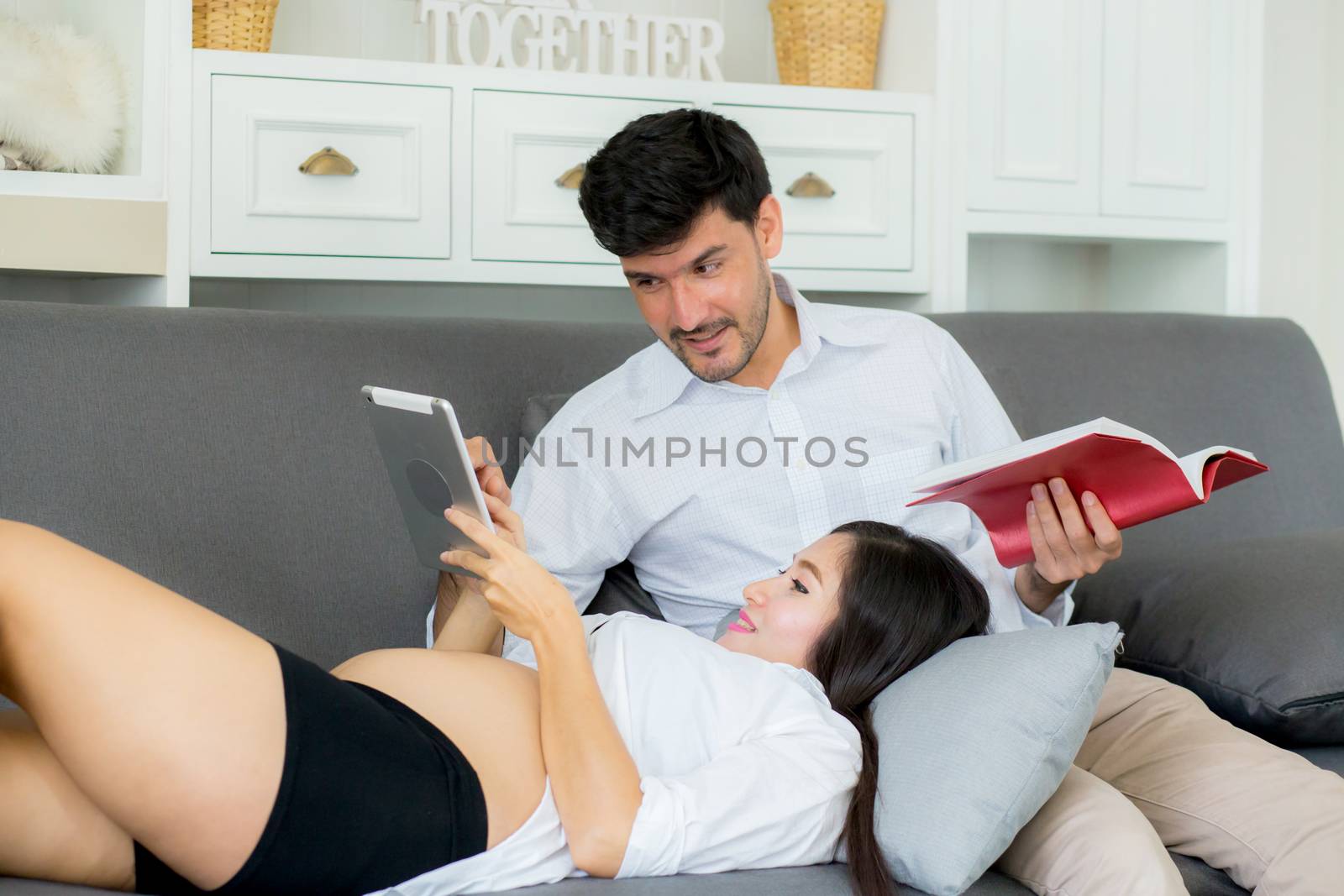 Asian couple family young mother and husband using a tablet at home, woman and man is pregnant lying on sofa looking tablet read book.