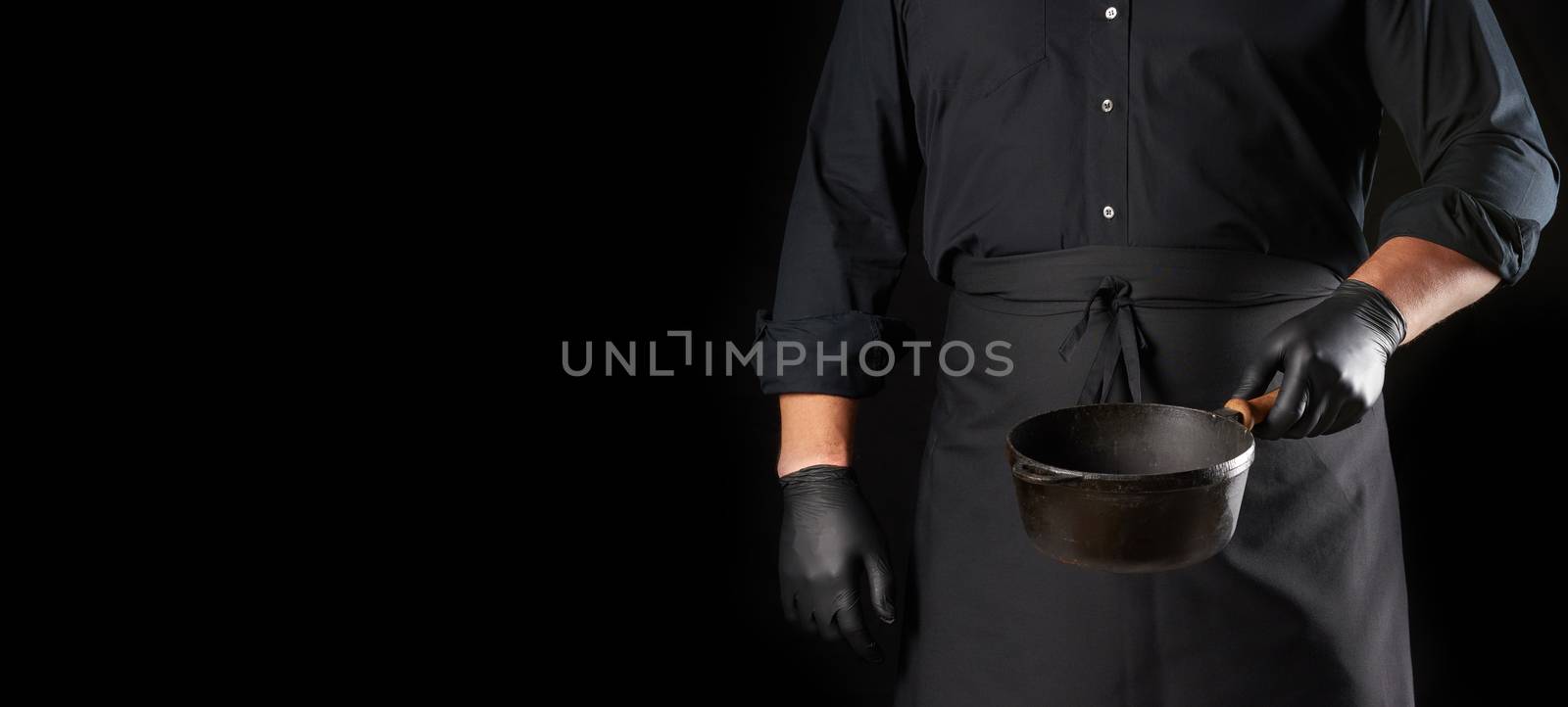 cook in black uniform and latex gloves holds an empty round vintage black cast iron pan in front of him, copy space