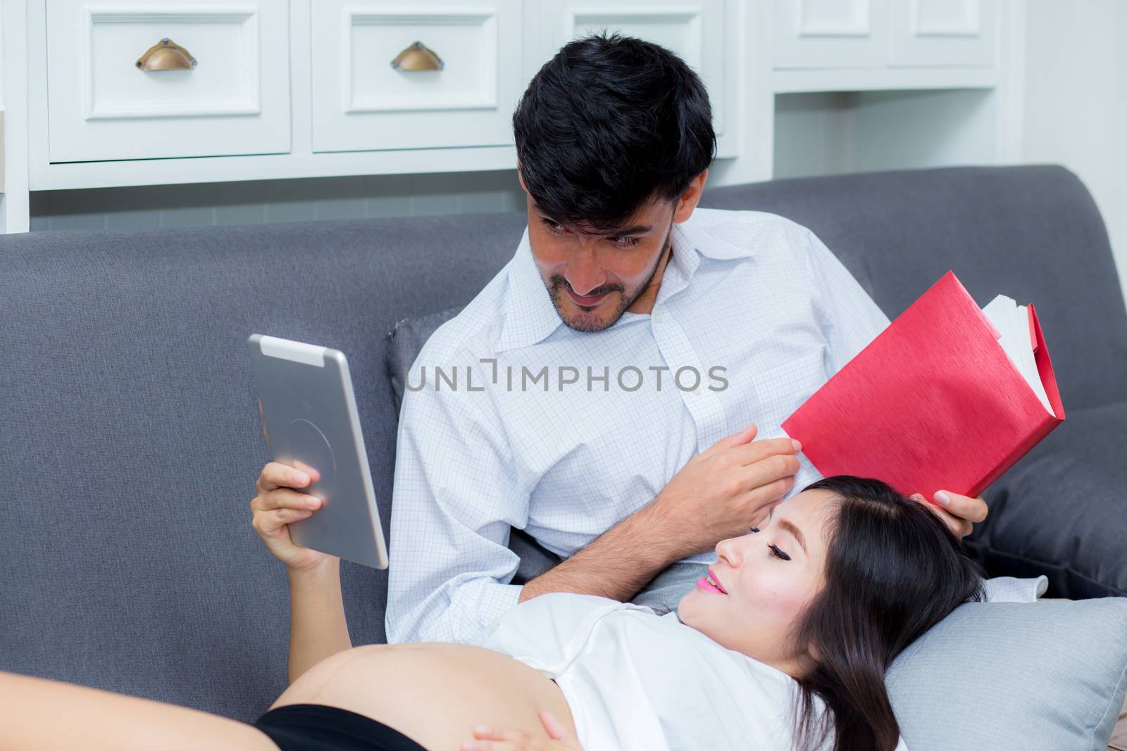 Asian couple family young mother and husband using a tablet at home, woman and man is pregnant lying on sofa looking tablet read book.