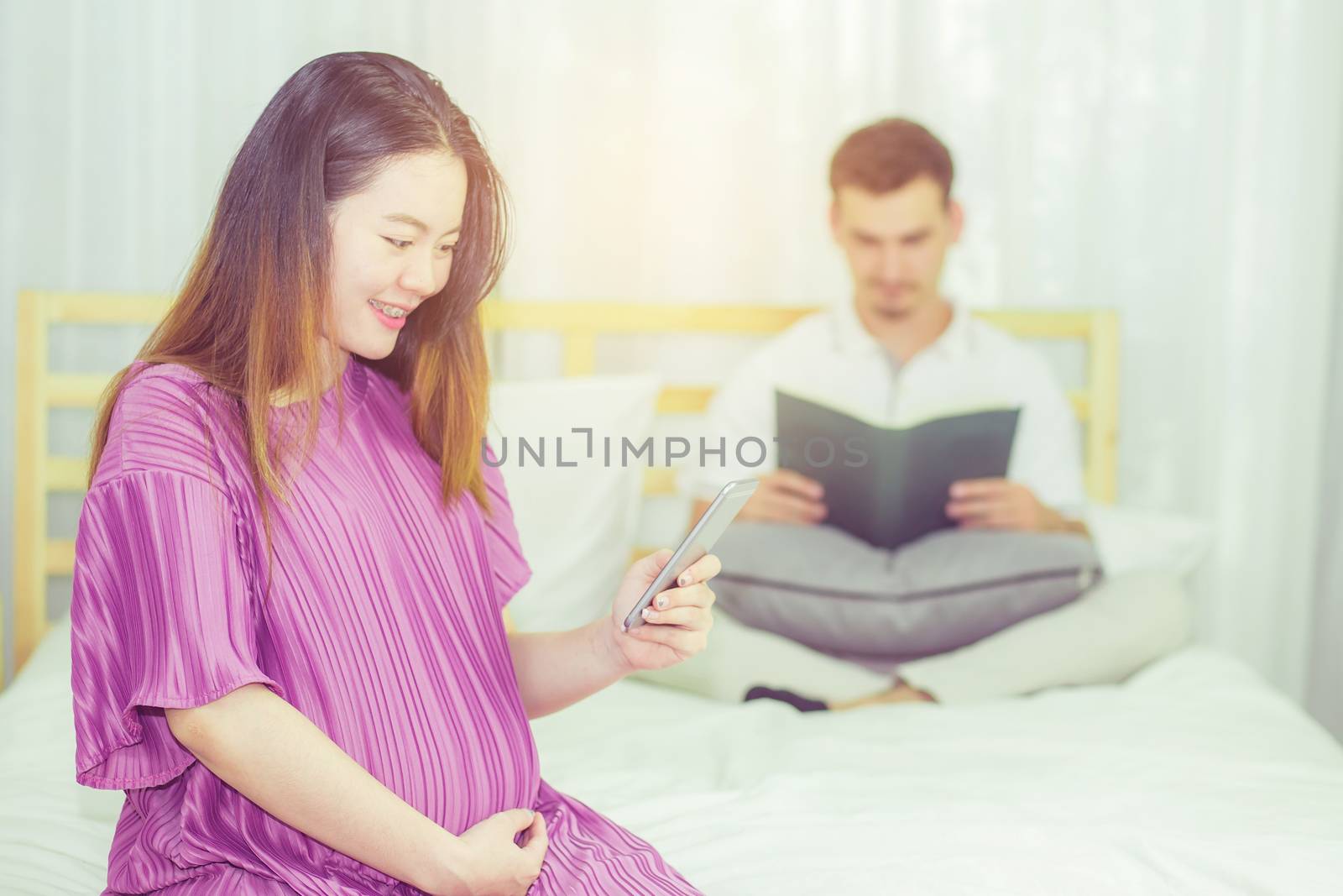 Asian pregnant people and expectation concept - happy pregnant woman holding mobile phone sitting on bed and touching belly at home with father reading book background.