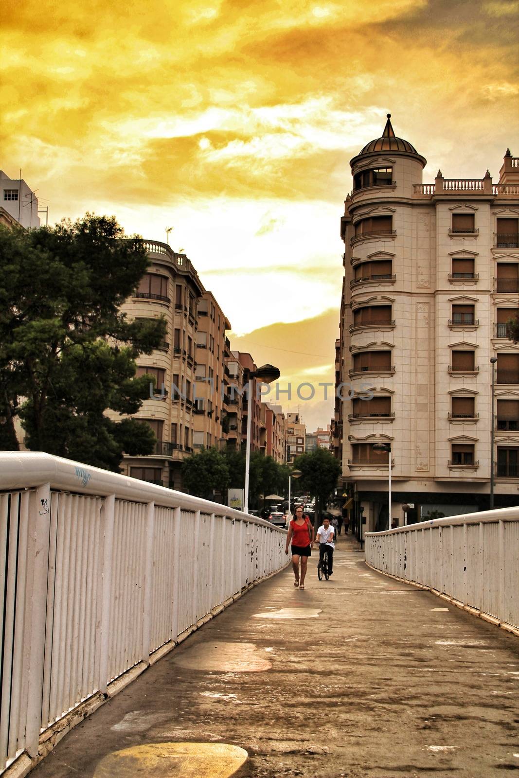 Elche, Alicante, Spain- September 12, 2018: People crossing a bridge at sunset in Elche. Beautiful buildings in the background.