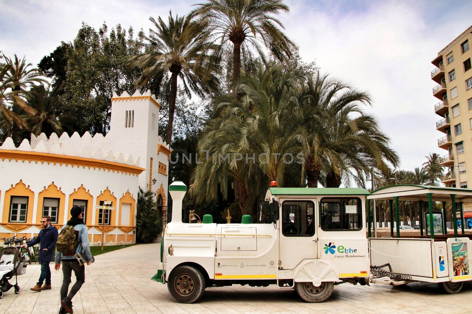 Elche, Alicante, Spain- February 19, 2019: Guided tour on Tourist train through the streets and palm groves in winter under blue sky. Tourist Office in the background.