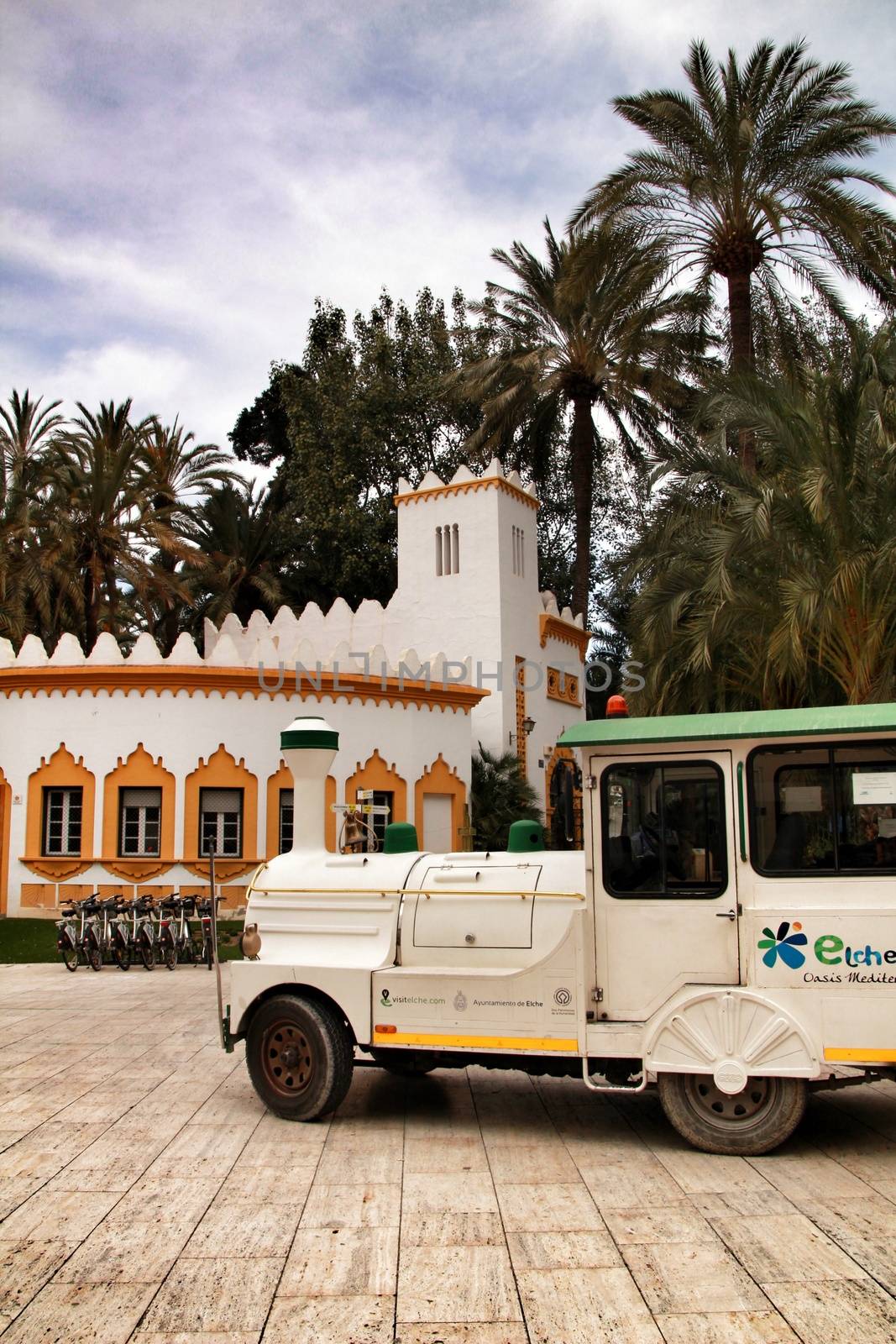 Elche, Alicante, Spain- February 19, 2019: Guided tour on Tourist train through the streets and palm groves in winter under blue sky. Tourist Office in the background.