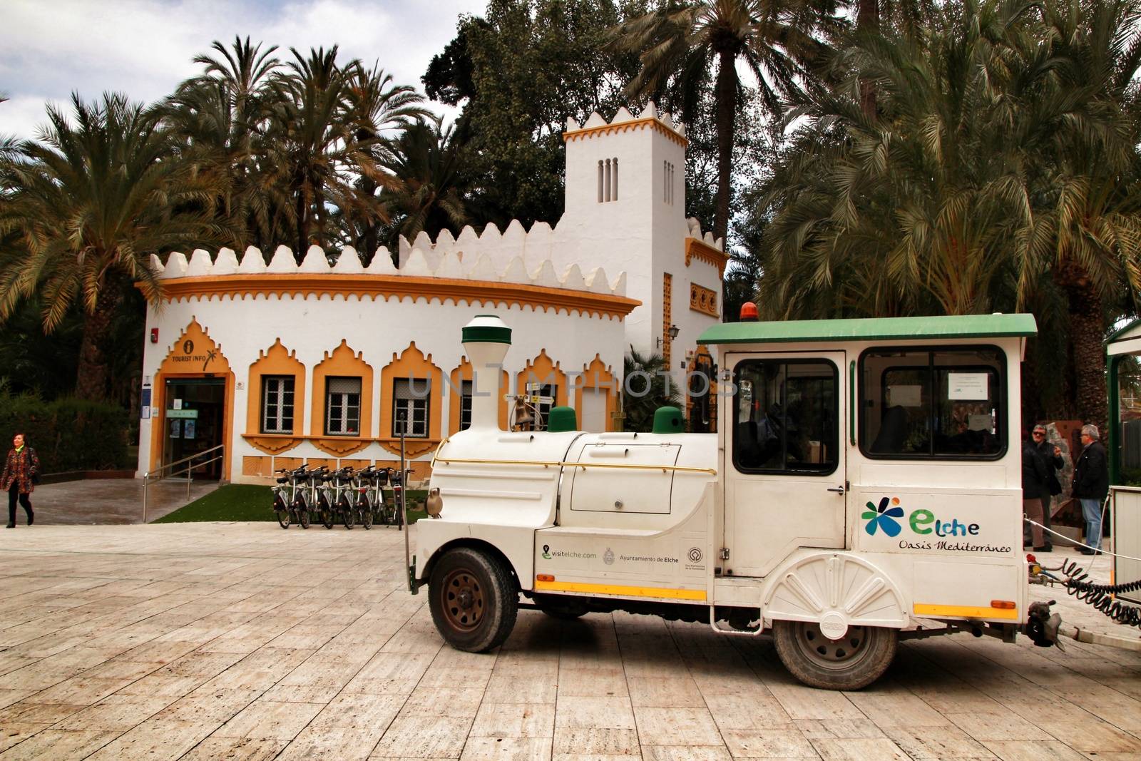 Elche, Alicante, Spain- February 19, 2019: Guided tour on Tourist train through the streets and palm groves in winter under blue sky. Tourist Office in the background.