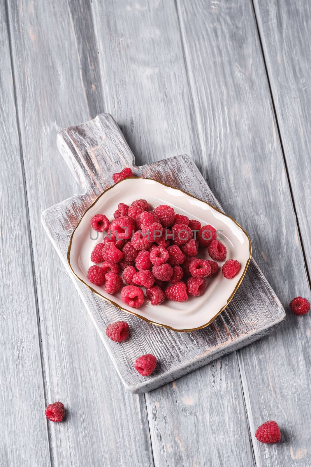 Raspberry fruits in plate on old cutting board, healthy pile of summer berries on grey wooden background, angle view