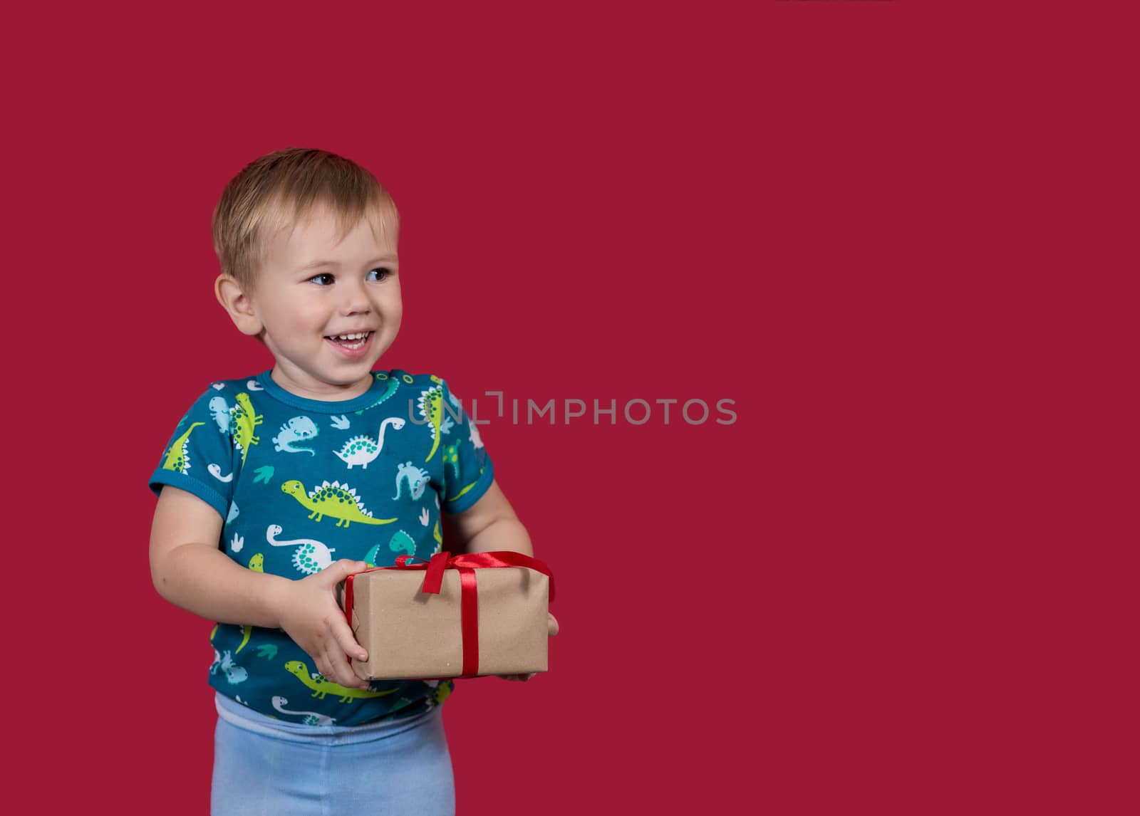 A little boy tries to unpack a New Year's gift with enthusiasm and excitement looking aside smiling on a red background