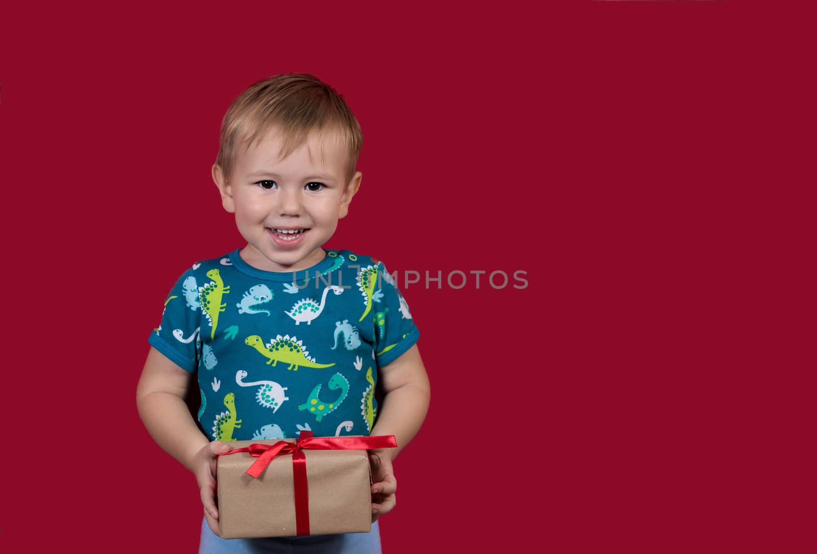 A little boy tries to unpack a New Year's gift with enthusiasm and excitement looking at the camera smiling on a red background