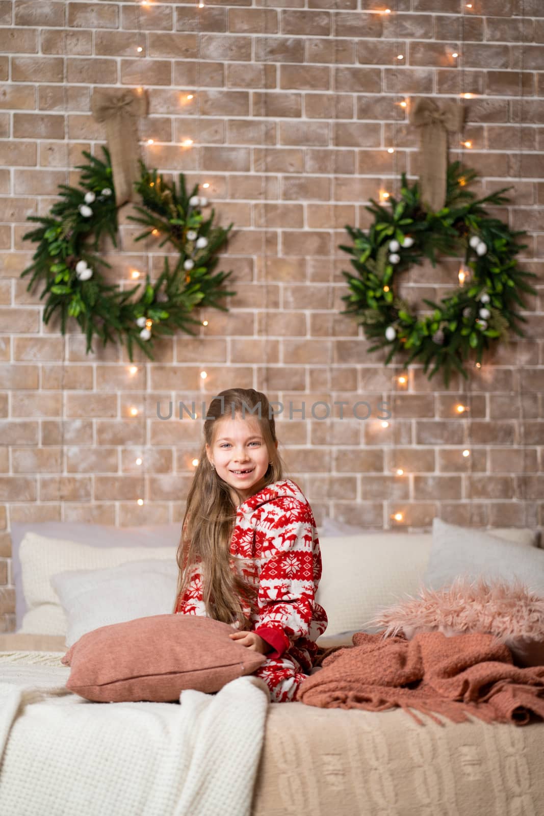 Little girl in red pajamas on the bed in a cozy Christmas interior.