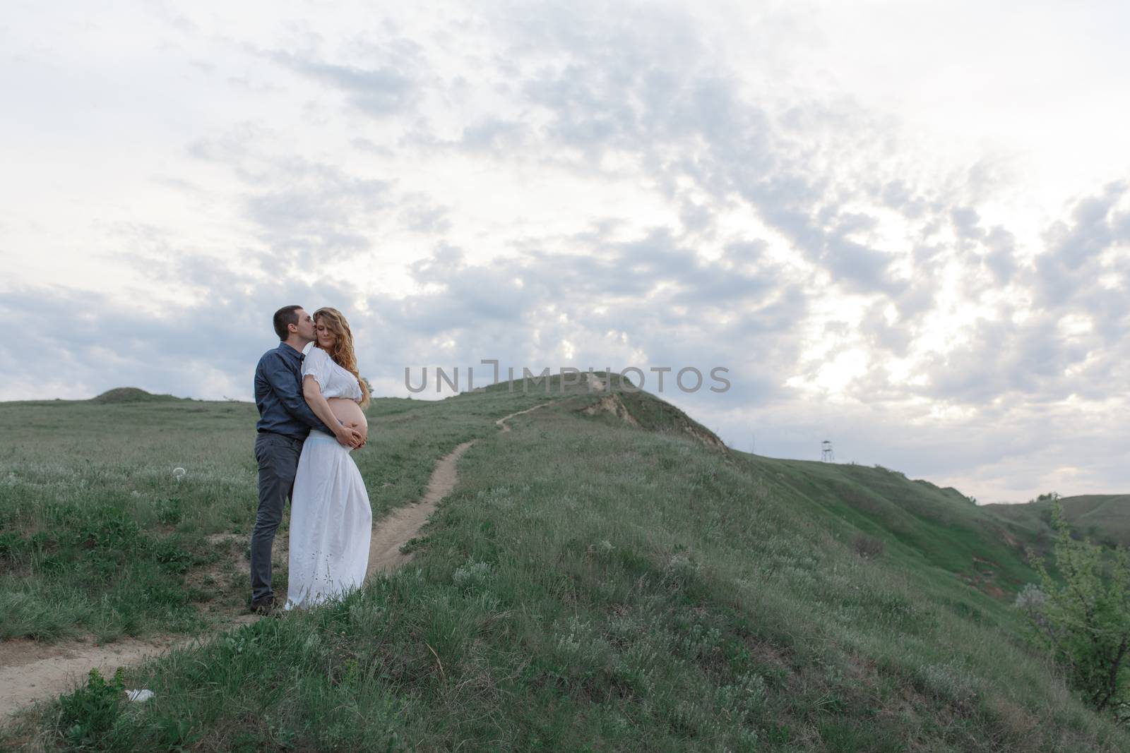 A couple in love, a pregnant wife in a white outfit are walking in a hilly field. Overcast weather by Try_my_best