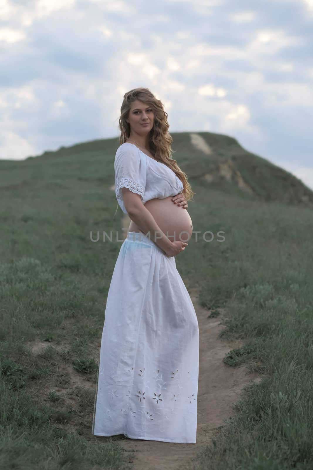 A pregnant girl in white is waiting for her beloved stand by a hill with a beaten track. Rural natural landscape.