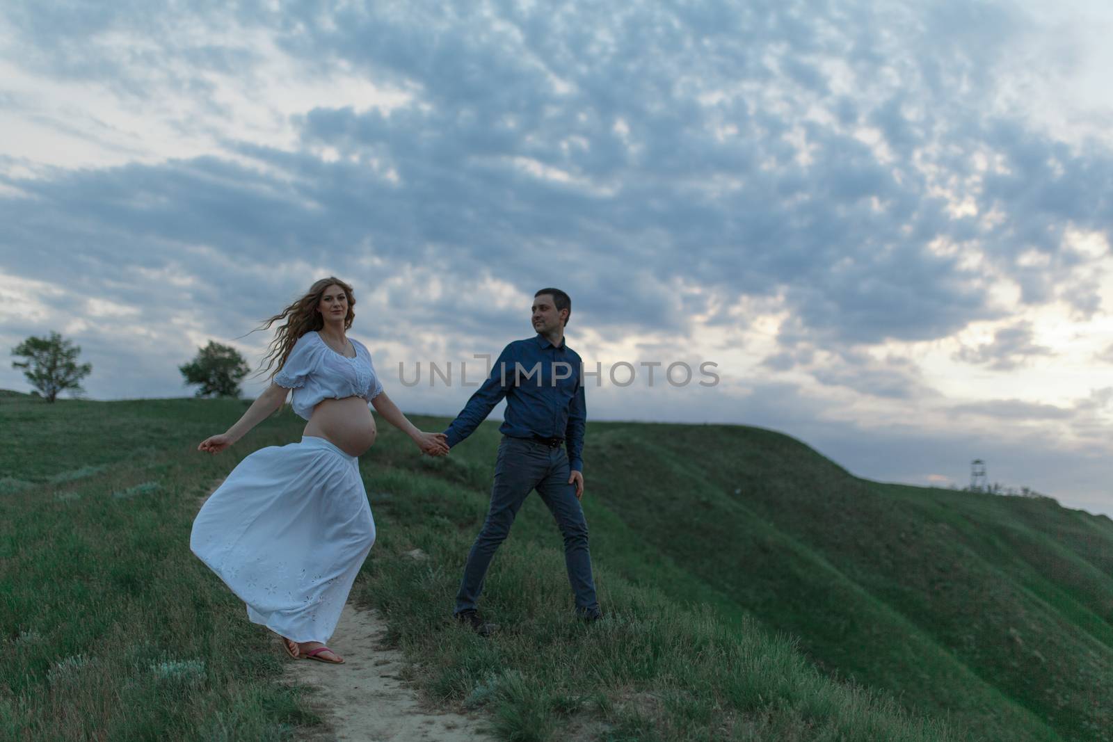 A couple in love, a pregnant wife in a white outfit are walking in a hilly field. Overcast weather by Try_my_best