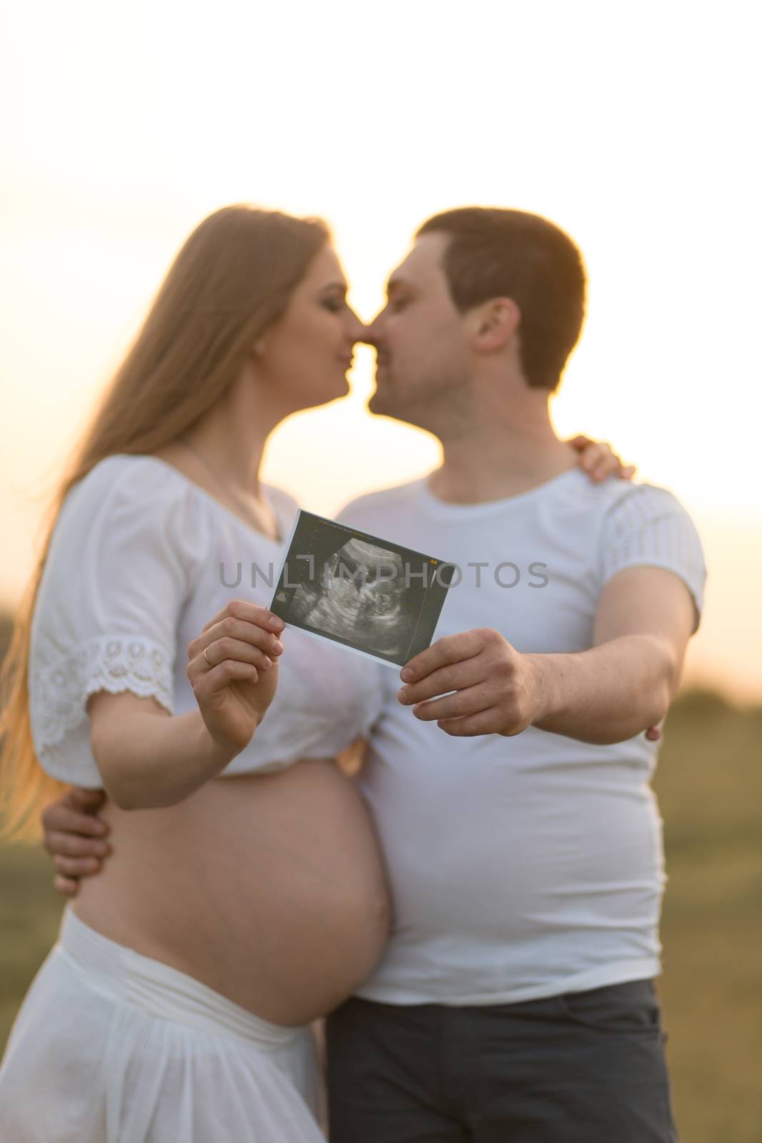Young couple kissing at sunset, holding an ultrasound scan with a baby. The girl is pregnant.