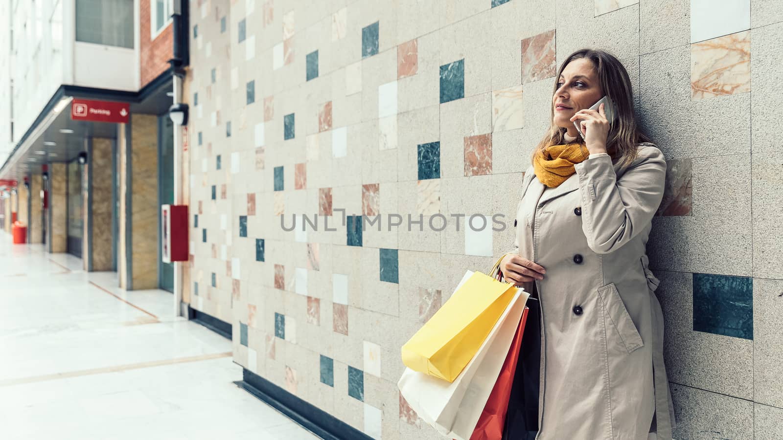 Adult woman wearing a beige raincoat and yellow scarf talking on her mobile phone while holding shopping colorful bags. Space for your text.