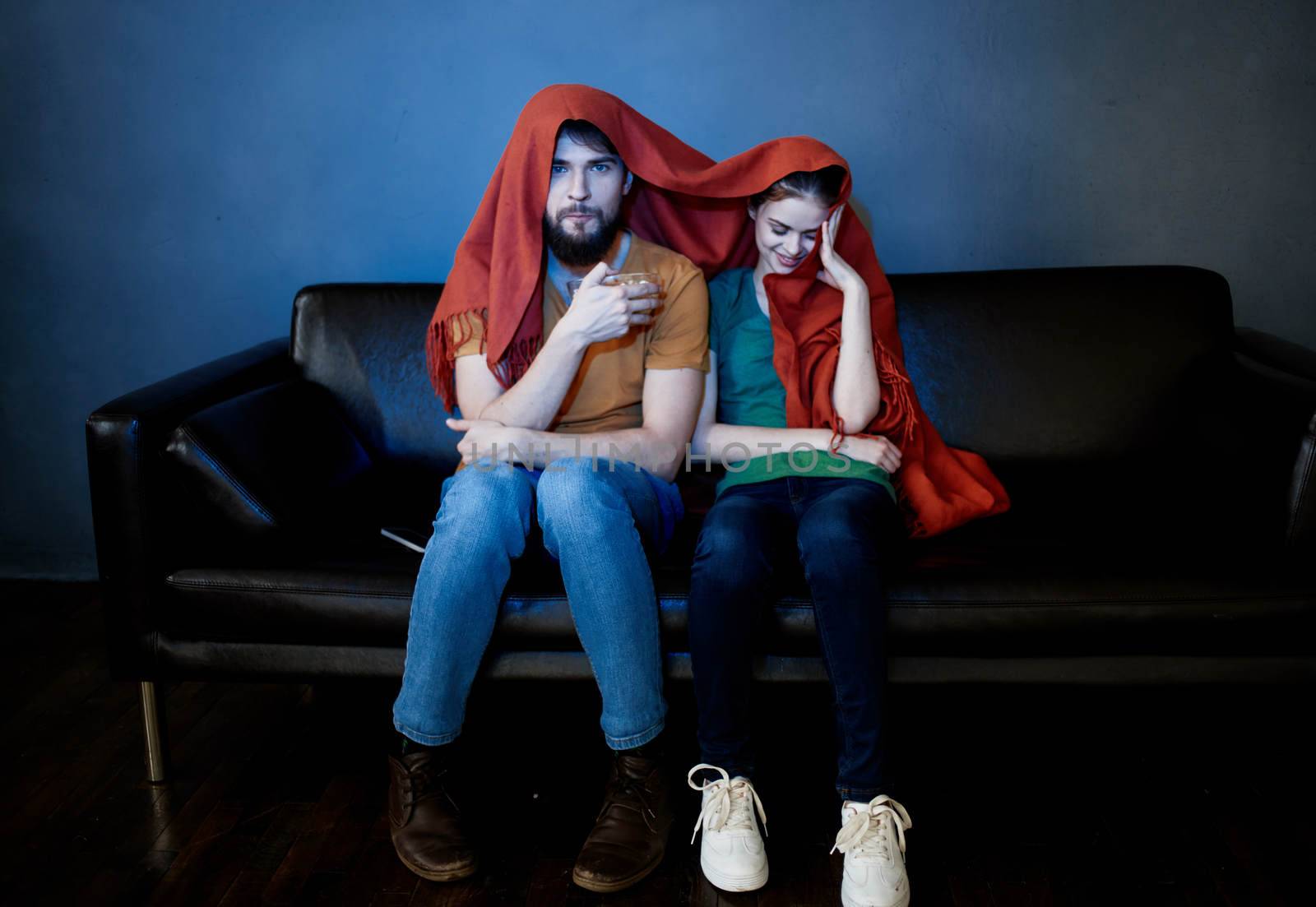 a woman and a man under a red blanket on the couch watching tv in the evening. High quality photo