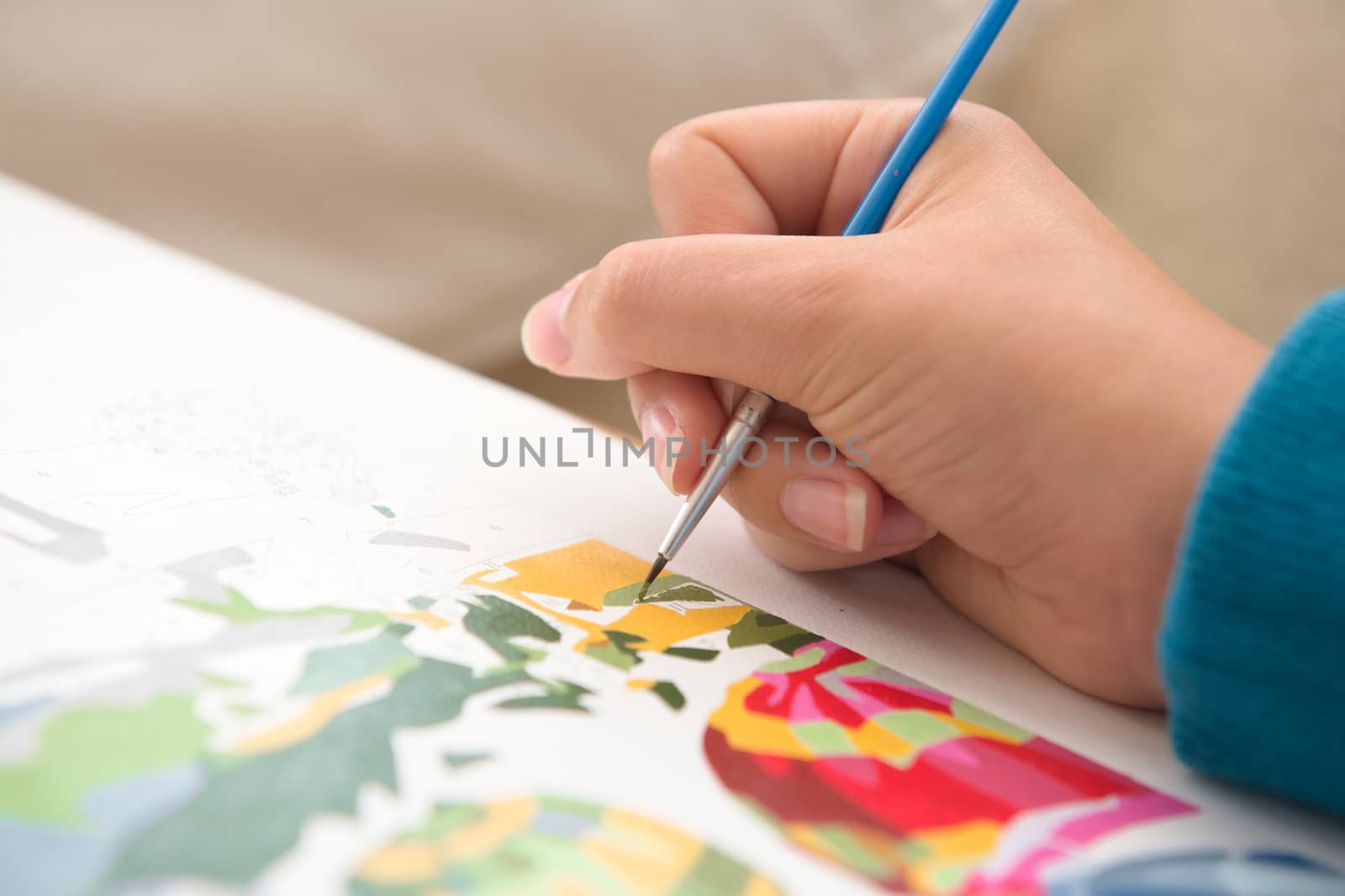 8 - Detailed close up of thin paint brush being used by a young female to paint by numbers. Multiple colors and vivid blues contrast the warm skin.