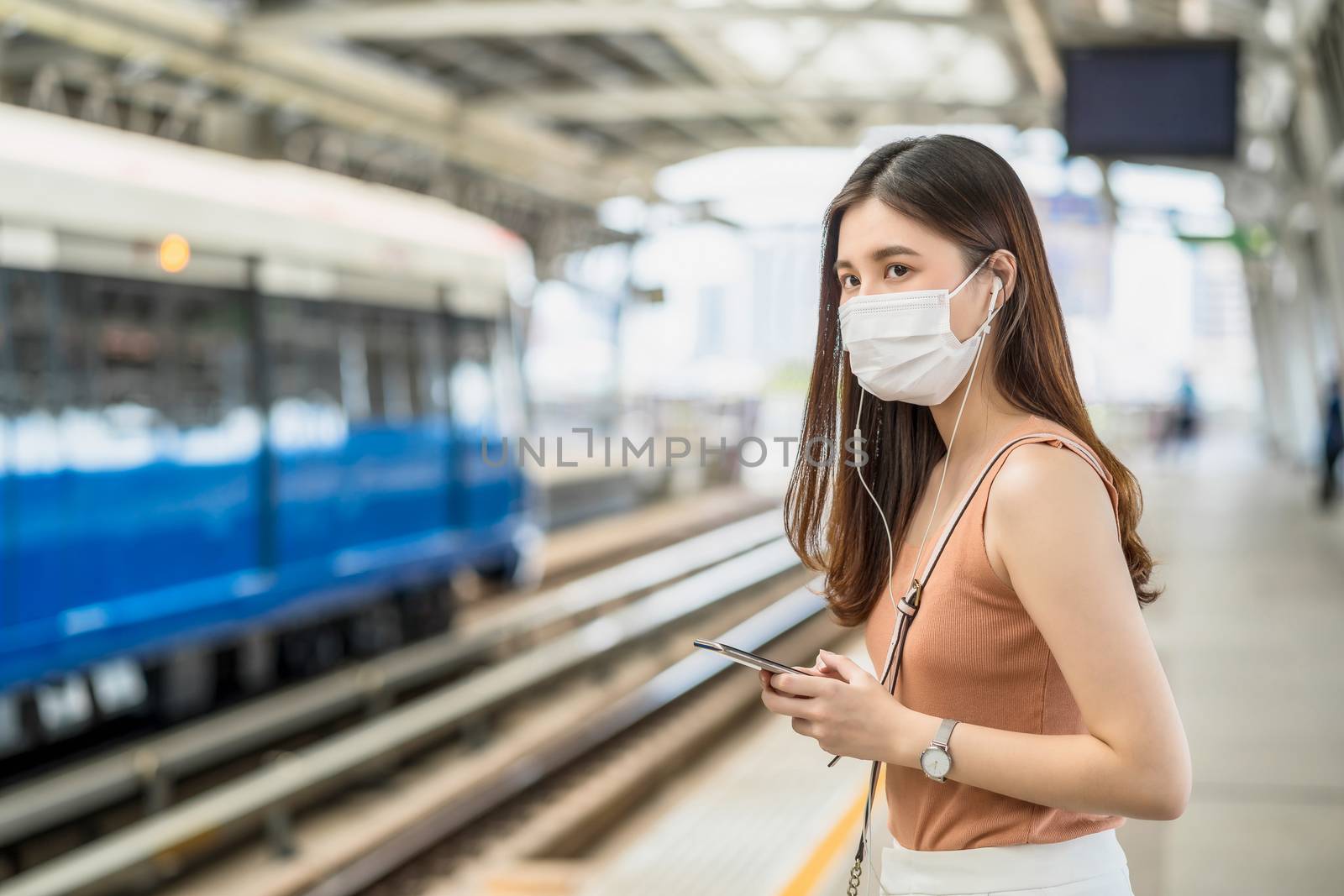 Young Asian woman passenger wearing surgical mask and listening music via smart mobile phone in subway train when traveling in big city at Covid19 outbreak, Infection and Pandemic concept