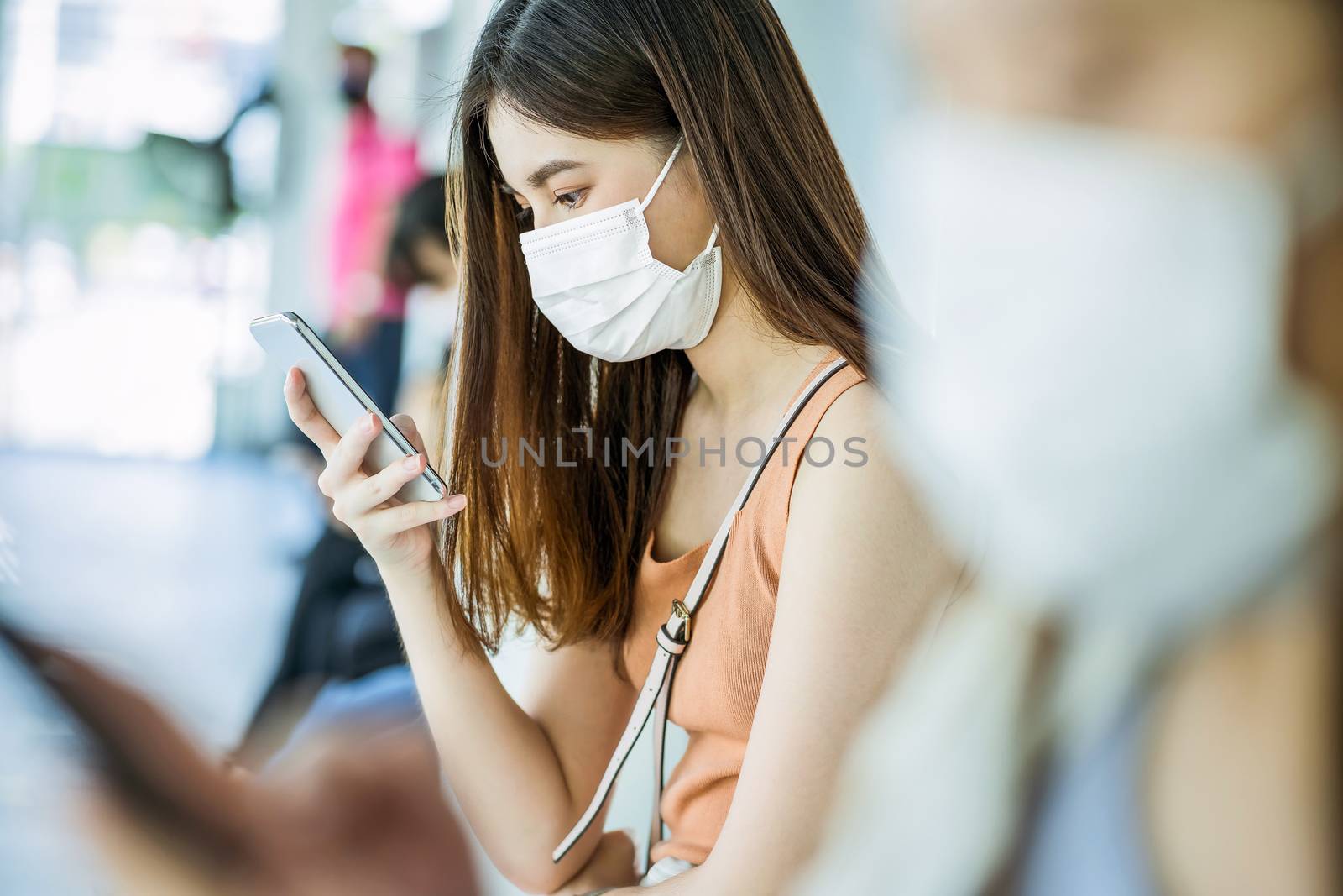 Young Asian woman passenger wearing surgical mask and using mobile phone for social network when waiting bus at station with crowned people when traveling in big city at Covid outbreak, Infection and Pandemic concept