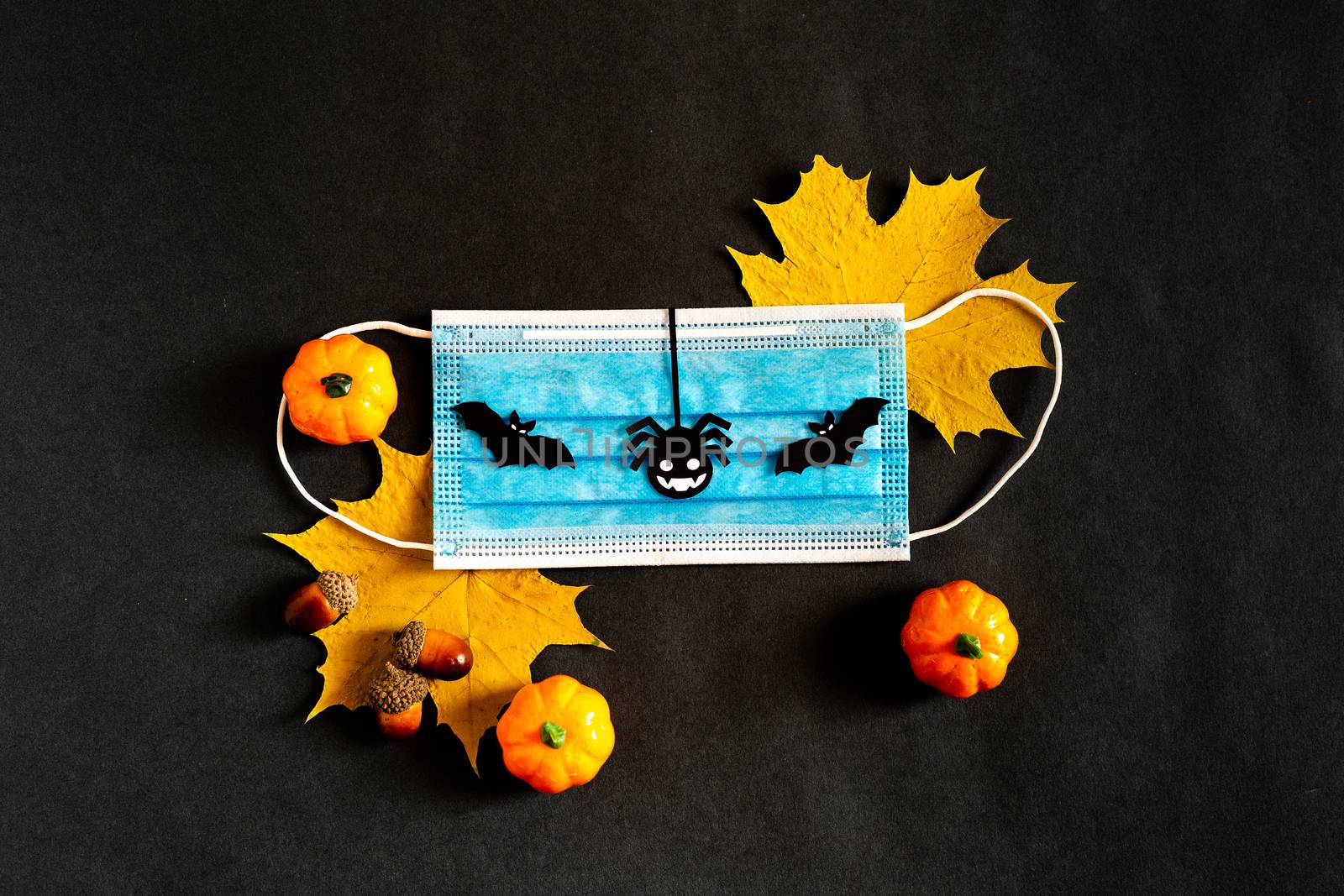 Halloween during the pandemic. Copy space. The view from the top. In the fall of 2020. Yellow maple leaves with a medical mask on a black background with black paper silhouettes, bat, pumpkin, spider by Pirlik