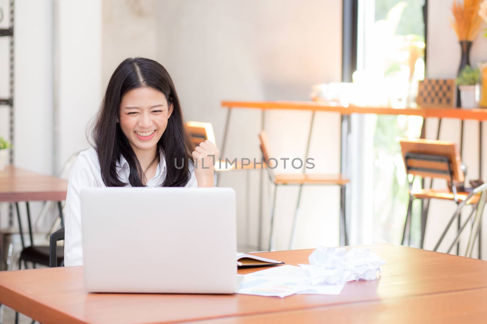 Beautiful asian young woman working online on laptop sitting at coffee shop, professional female freelancer with paper crumpled and using notebook computer with connect to internet for distance job.