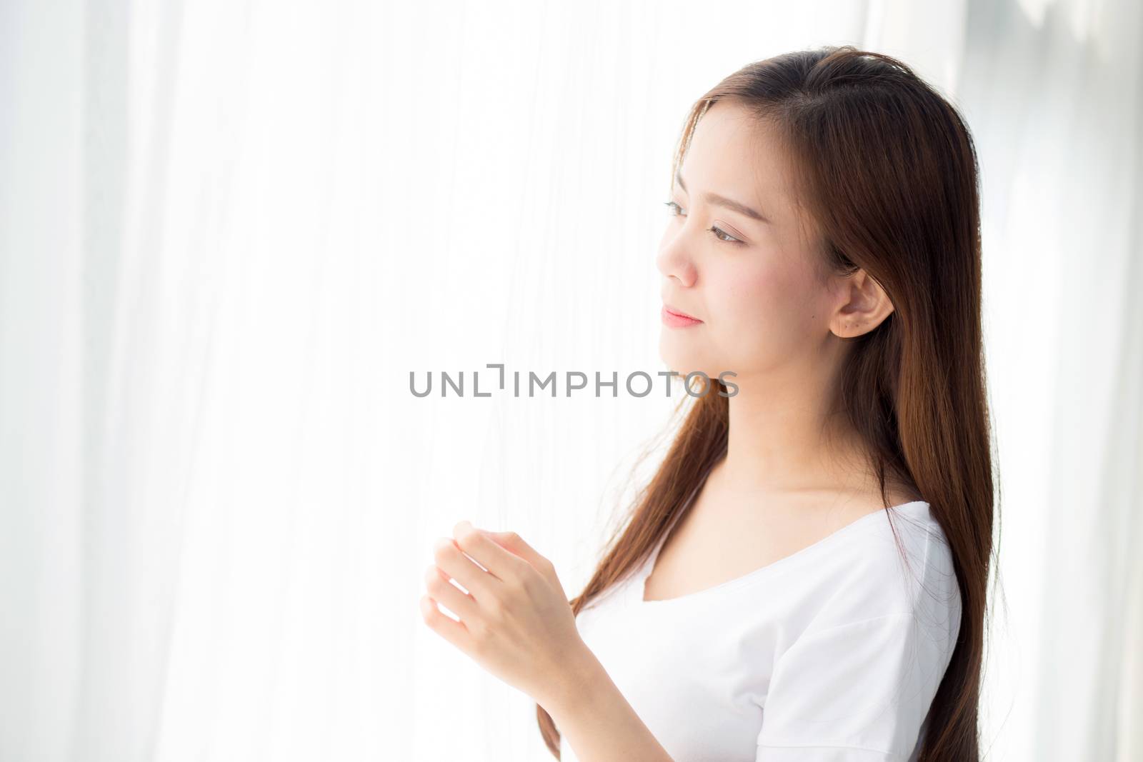 Beautiful young asian woman standing the window and smile while wake up with sunrise at morning, lifestyle and relax concept.