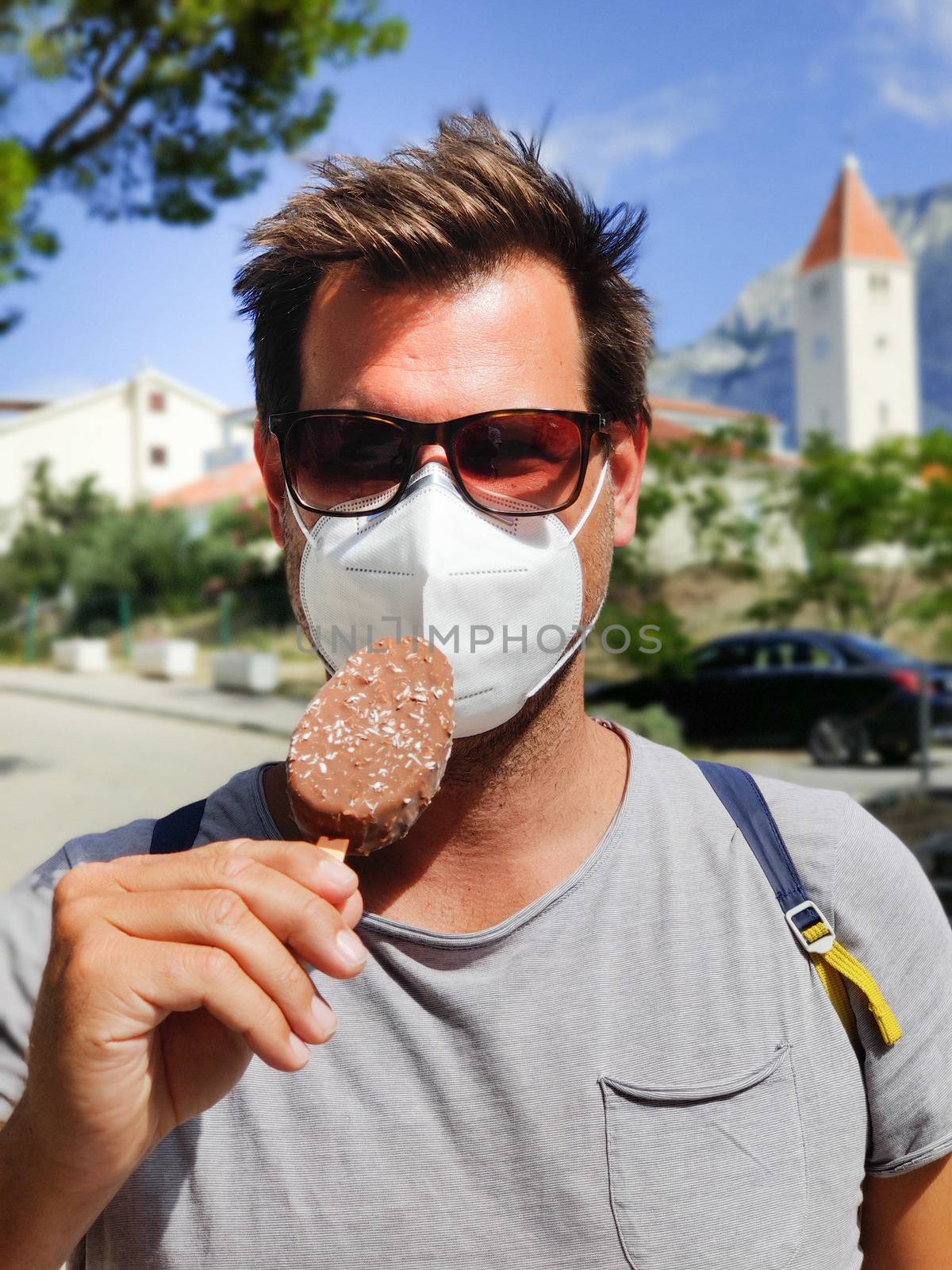 Young guy on summer vacations wearing corona virus protective face mask not beeing able to lick ice cream bar by kasto