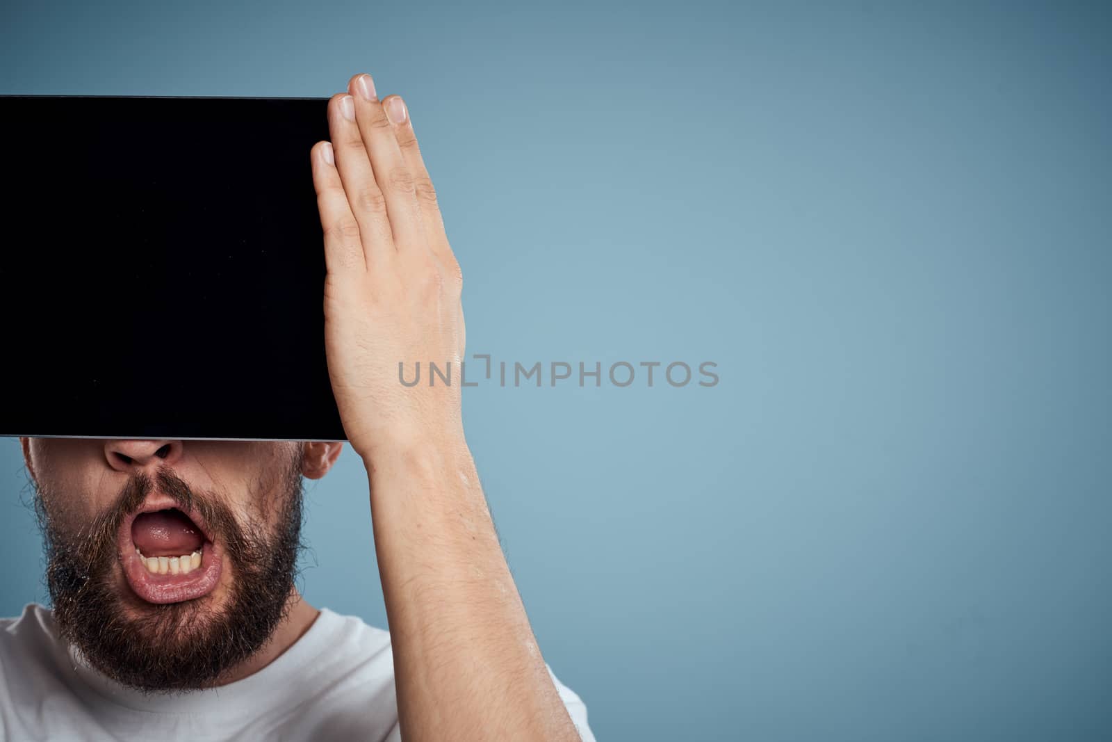 man with touch tablet in front of face on blue background cropped view copy space emotions model by SHOTPRIME