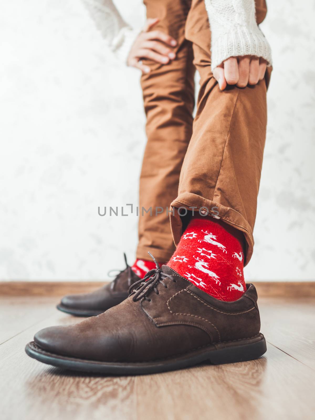 Young man pulls up leg of his chinos trousers to show bright red socks with reindeers on them. Scandinavian pattern. Winter holiday spirit. Casual outfit for New Year and Christmas celebration. by aksenovko