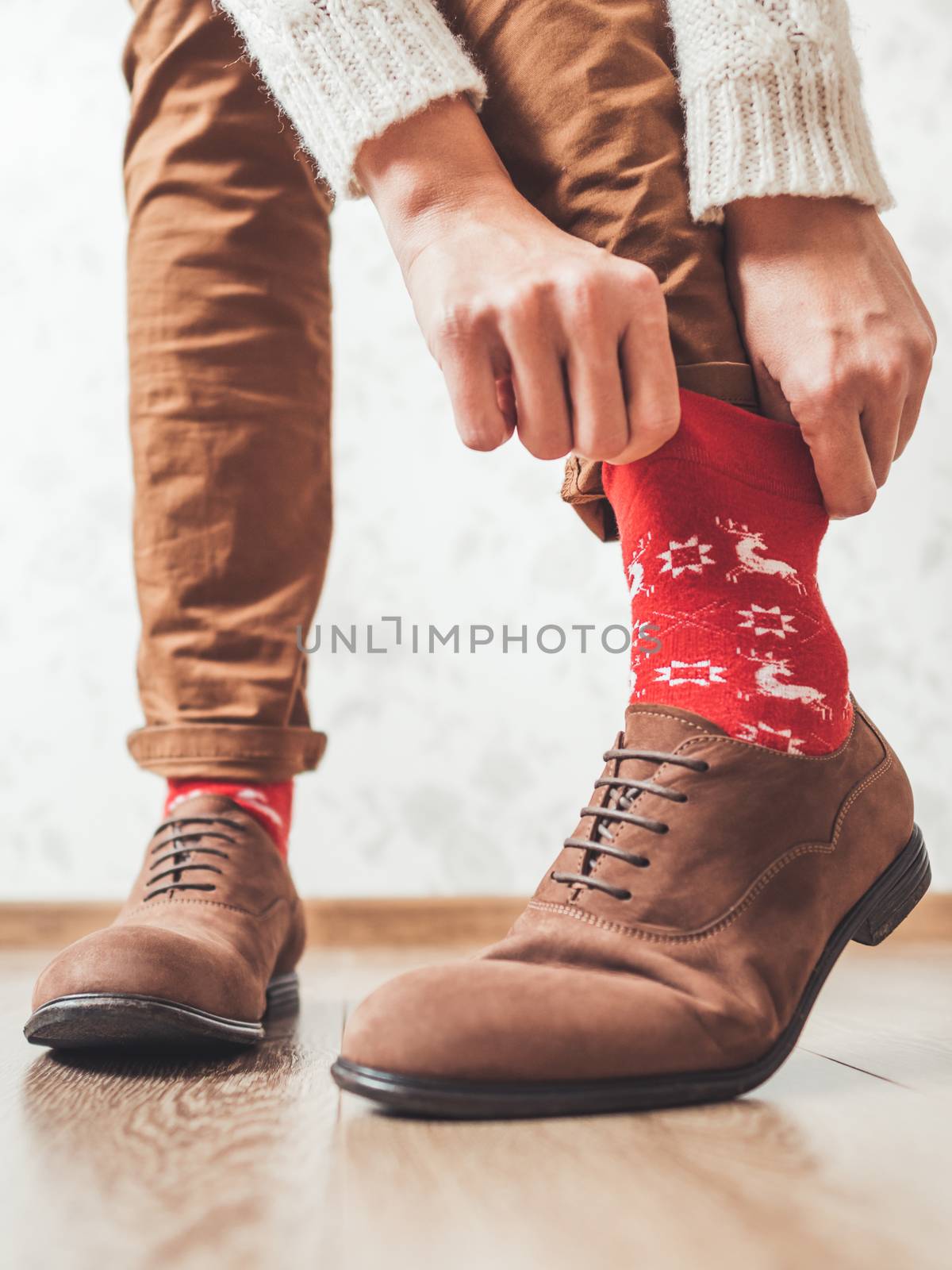 Young man pulls up leg of his chinos trousers to show bright red socks with reindeers on them. Scandinavian pattern. Winter holiday spirit. Casual outfit for New Year and Christmas celebration. by aksenovko