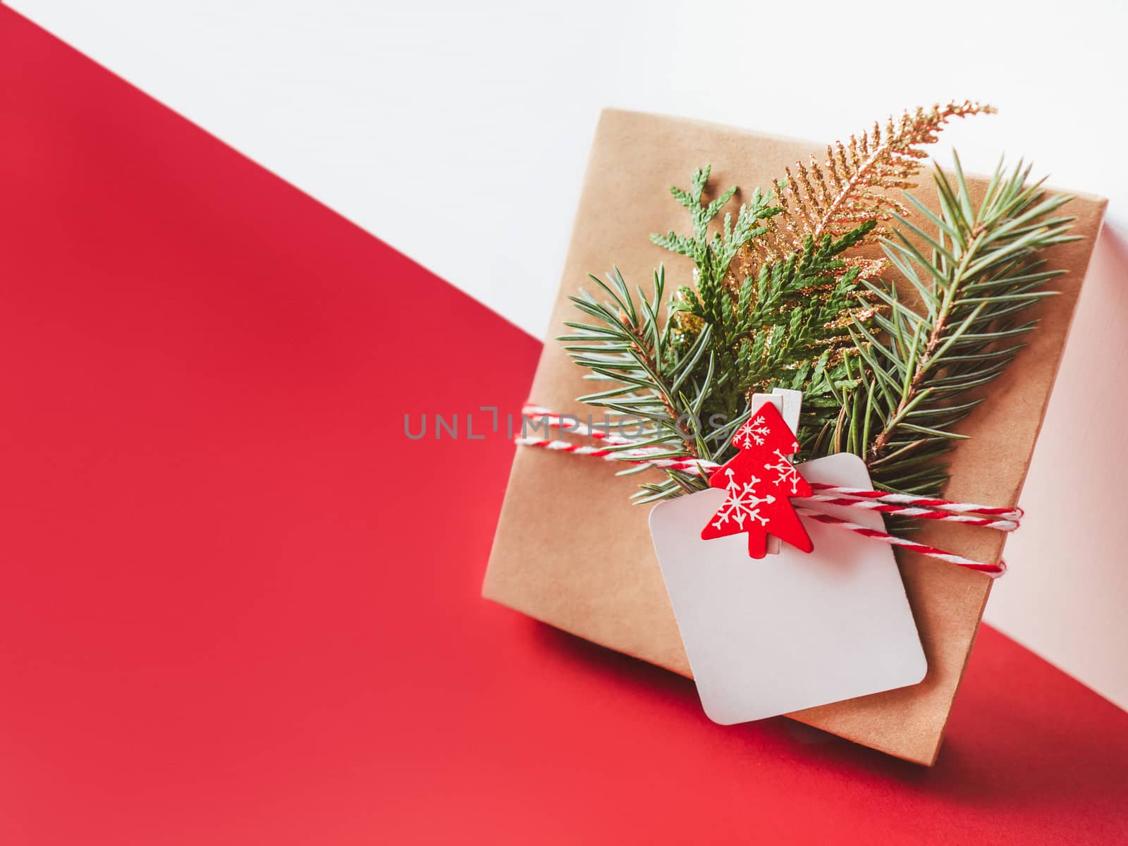 New Year present on geometrical white and red festive background. Gift wrapped in craft paper with fir tree twigs, red Christmas tree and clear tag. Copy space on label. Winter holiday spirit.