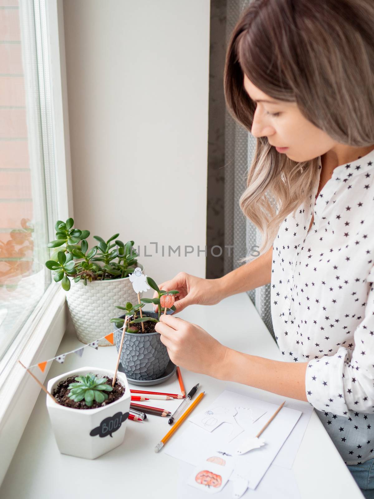 Woman decorates flower pots with handmade decorations for Halloween. Painted ghost in medical protective mask and pumpkin in flower pot with succulent plant.