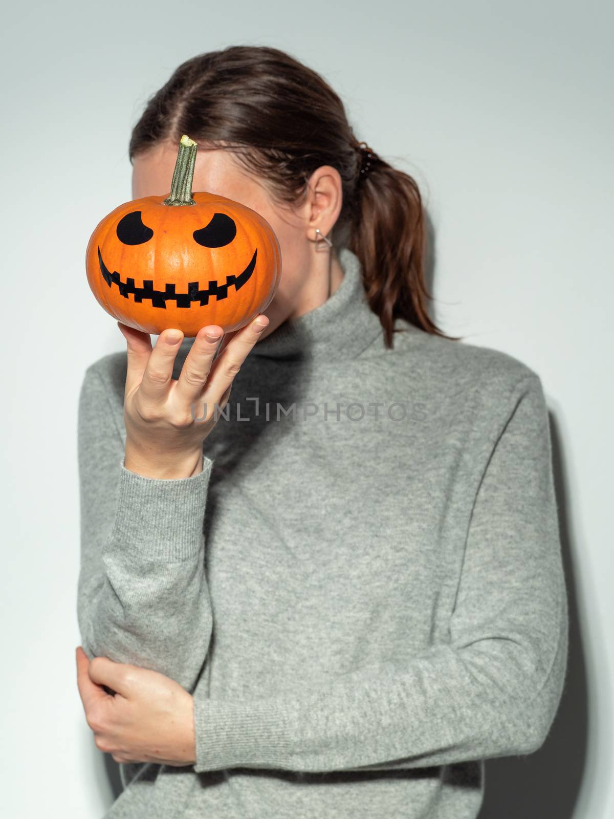 Crop unrecognizable woman holding halloween scary face bright orange pumpkin in front of her face. Young woman in gray cashemere sweater hides her face behind jack-o-lantern pumpkin. Snapshot style