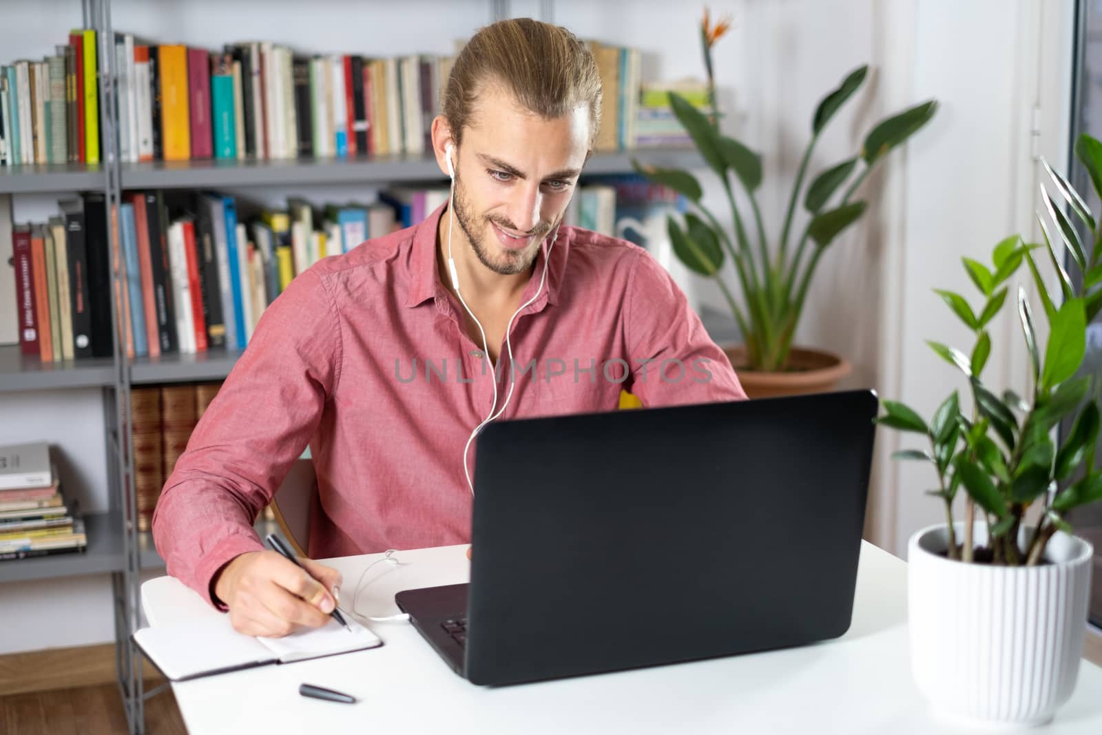 Attractive young man sitting at the table at home working with a laptop with happy face and smiling with a smile and writing on the book