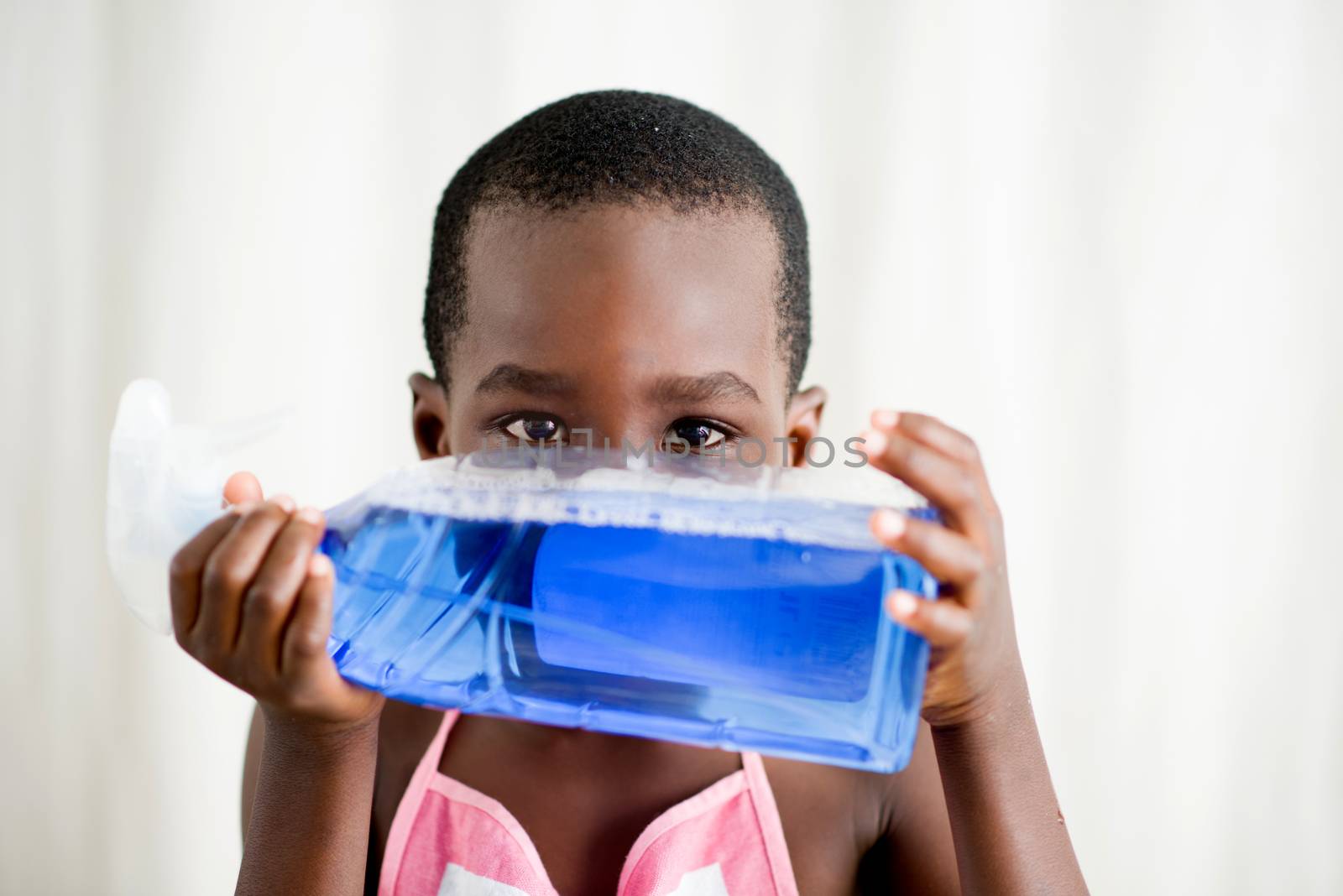 little boy in apron standing in studio hiding his face with a bottle of blue liquid soap