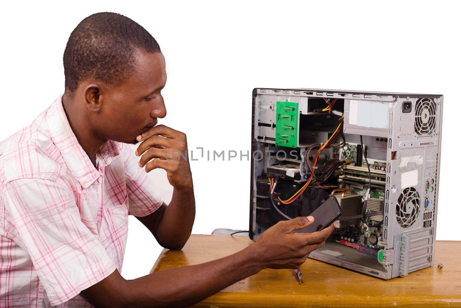 young technician sitting thinking with a desktop computer in front of him and holding a mobile phone.