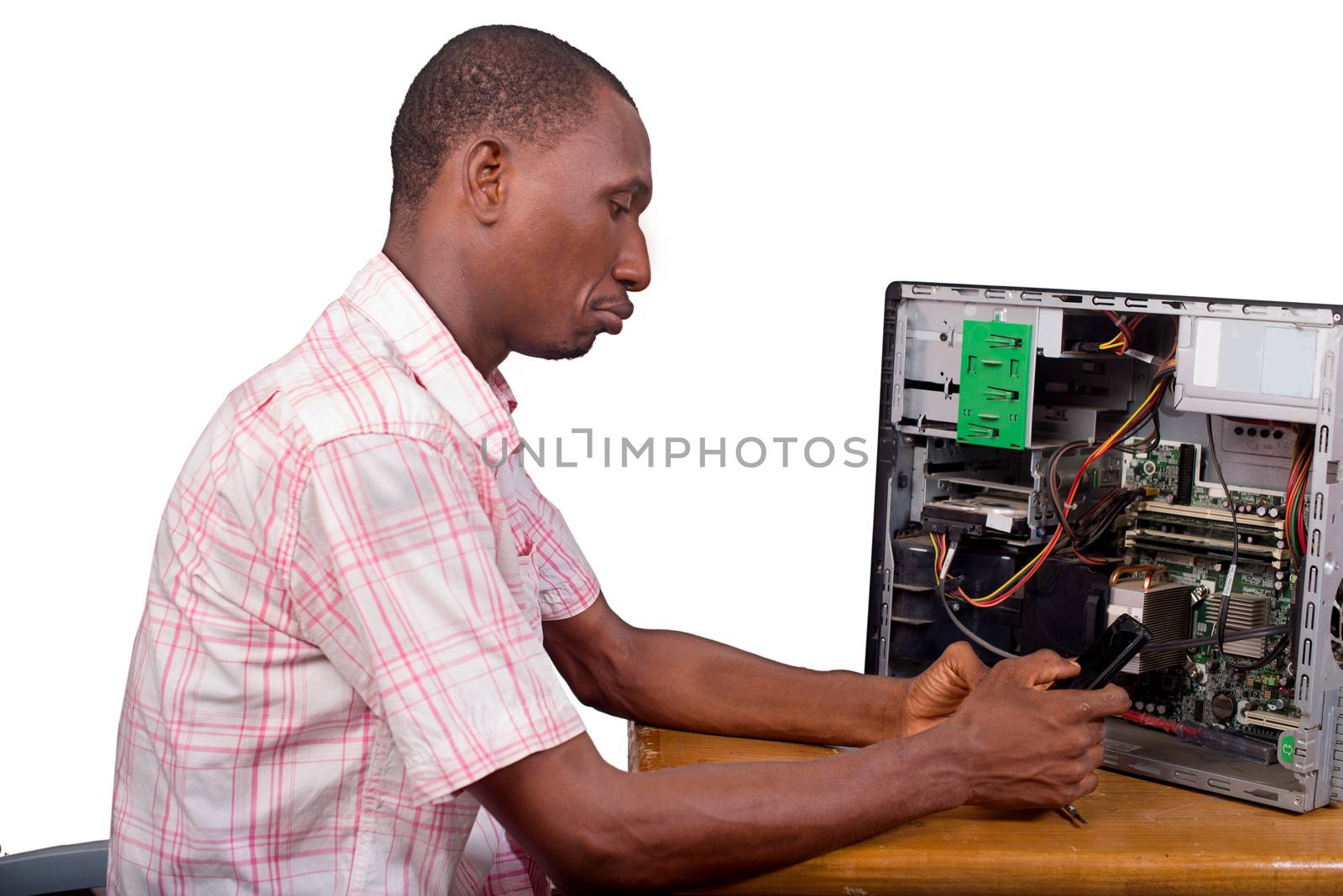 young technician sitting thinking with a desktop computer in front of him and holding a mobile phone.