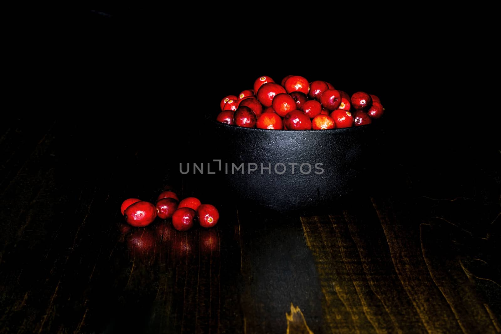 A bunch of cranberrries in a black, cast iron bowl, with black background and brown base. Shot in low key style.