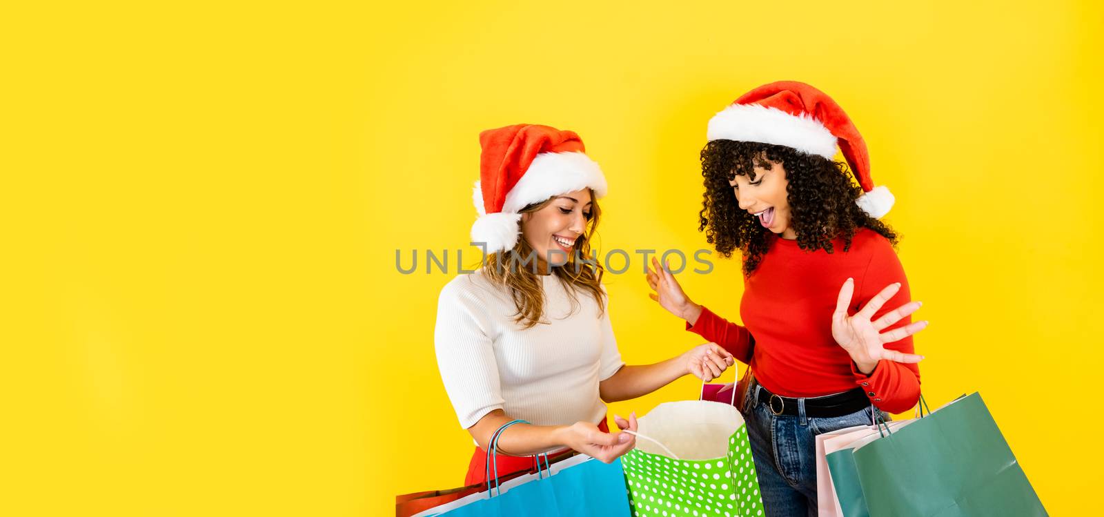 Cute Caucasian blonde young woman open her shopping bag showing to her black Hispanic female friend - Two millennial multiracial women shoppers with Santa hat and big yellow copy space studio shot by robbyfontanesi