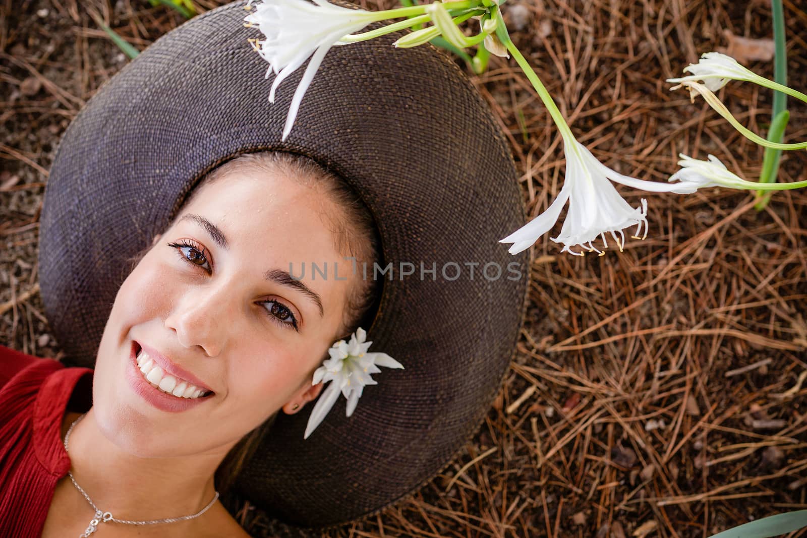 Closeup of a Beautiful caucasian young woman face lying on the ground on pine needles with a broad-brimmed dark hat, among sea daffodil (pancratium maritimum) flowers - Happiness living nature by robbyfontanesi