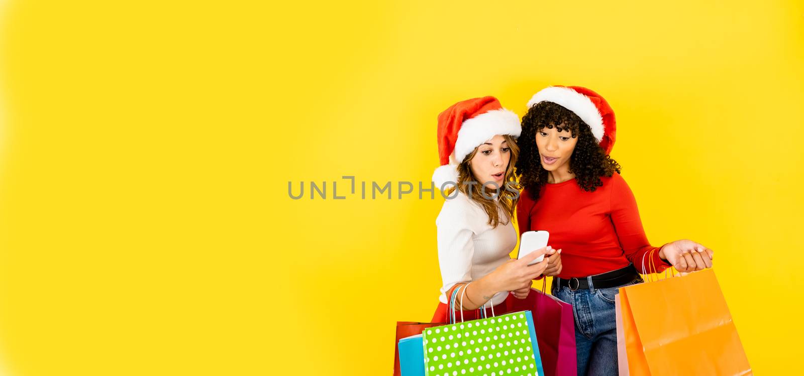 Two attractive women with Santa hat looking to a smartphone excited and surprised - Big yellow banner with female people doing shopping online holding colored shopping bags on yellow big copy space by robbyfontanesi