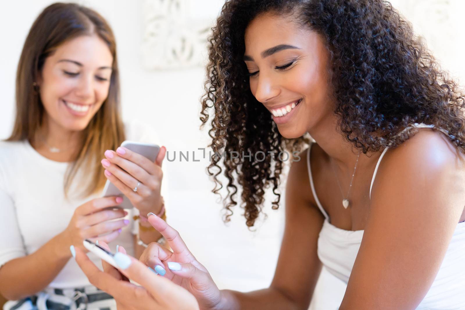 Two young beautiful women at home using smartphone - Cute brunette black Hispanic having fun indoor chatting on social networks with her beautiful blonde female friend - Focus on curly woman by robbyfontanesi