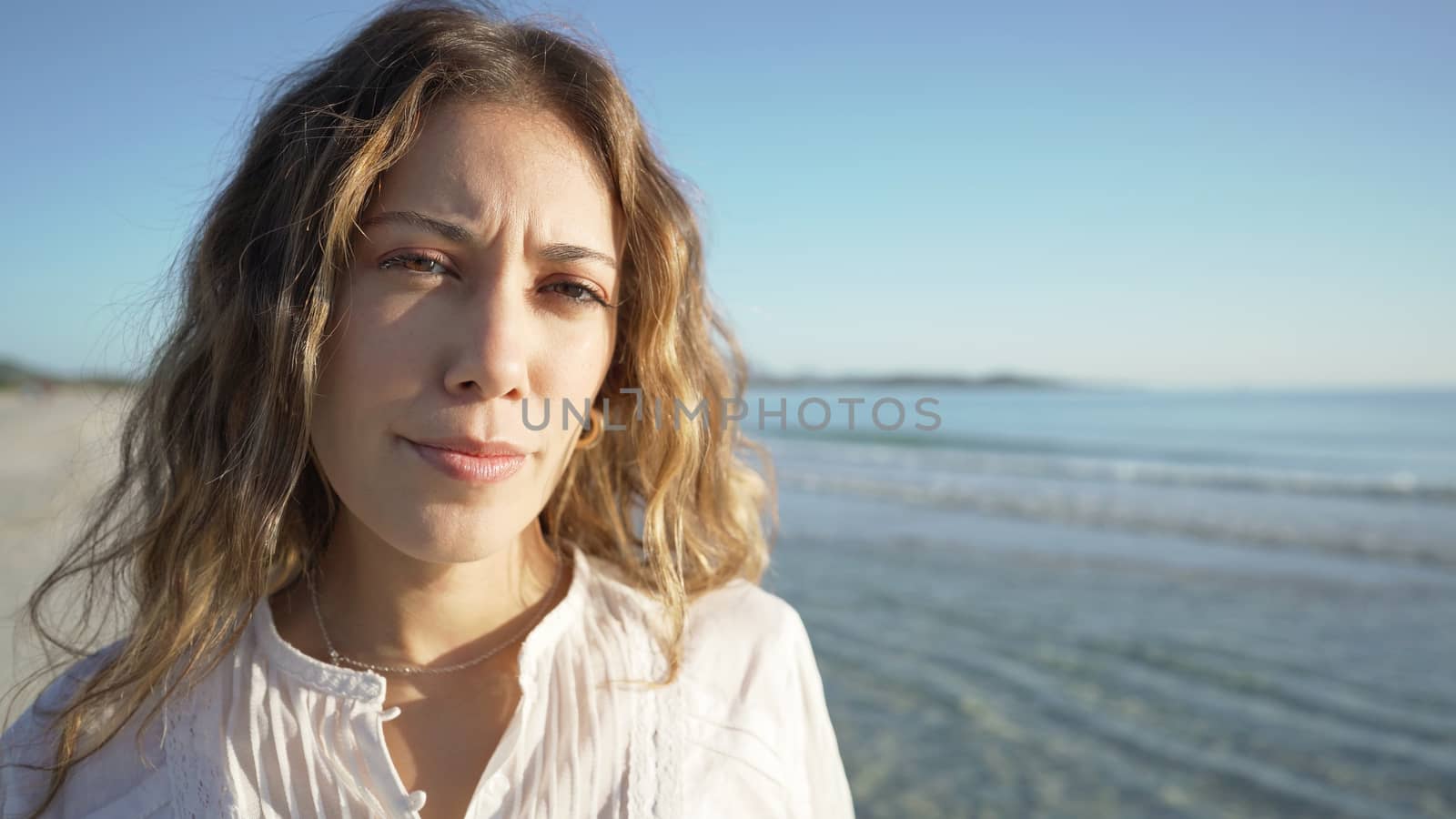 Face close-up of young cute Caucasian blonde woman looking at the camera at sunset or dawn in the seashore with a stylish white shirt - Real people confident expression